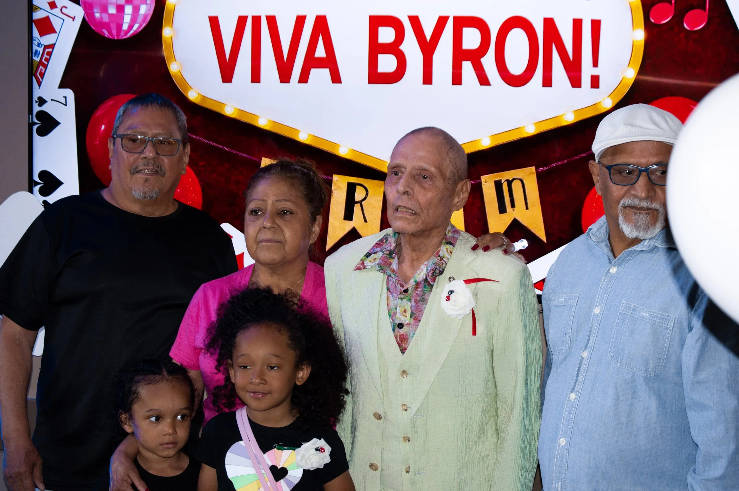 A group of people, including two children, standing in front of a sign that reads 'Viva Byron!' surrounded by decorative lights and balloons. The group appears to be at a celebration or event.