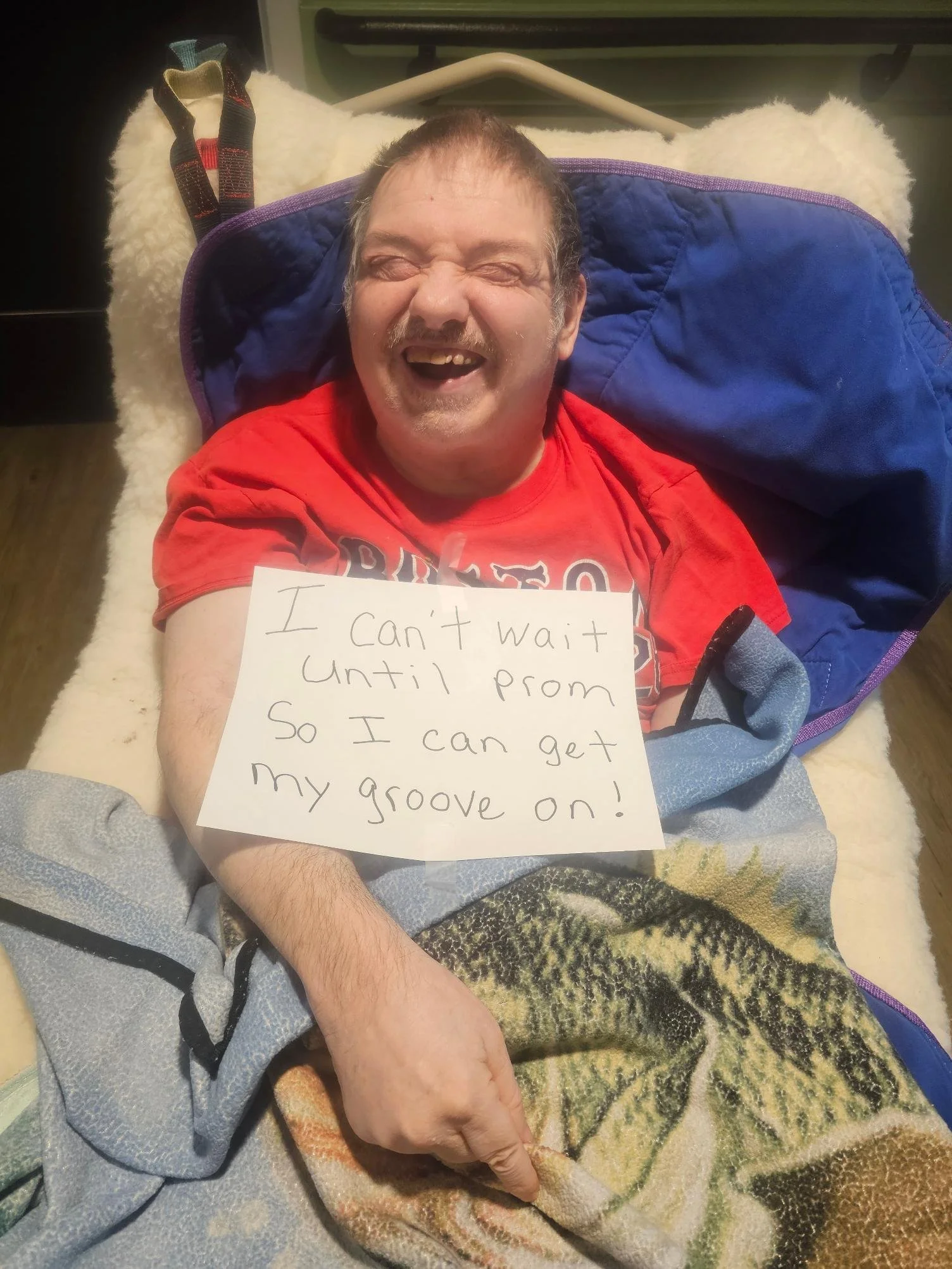A man is lying in a hospital bed, smiling widely, with a sign on his chest that reads, 'I can't wait until prom so I can get my groove on!'