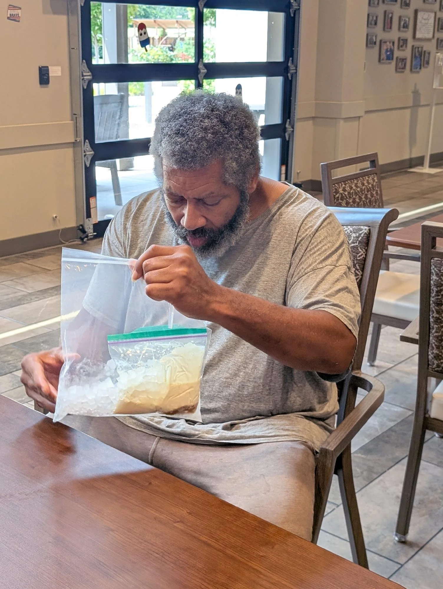 An elderly man with gray, curly hair and a beard is sitting at a wooden table, opening a clear plastic bag with a yellowish substance inside. He is wearing a gray T-shirt and is indoors, with a glass door and framed pictures on the wall behind him.
