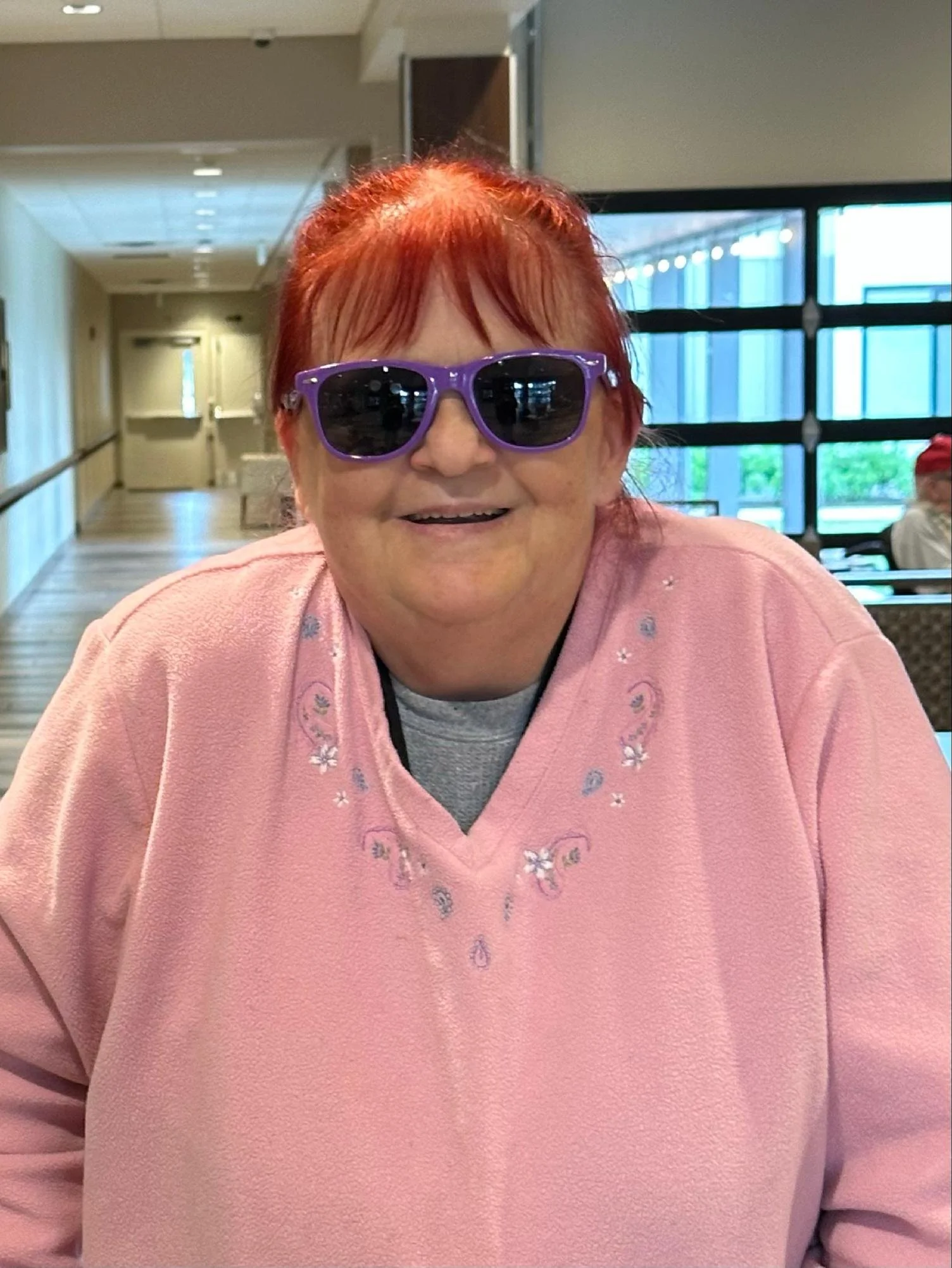 An elderly woman with red hair wearing purple sunglasses and a pink shirt with floral embroidery, sitting indoors near large windows.