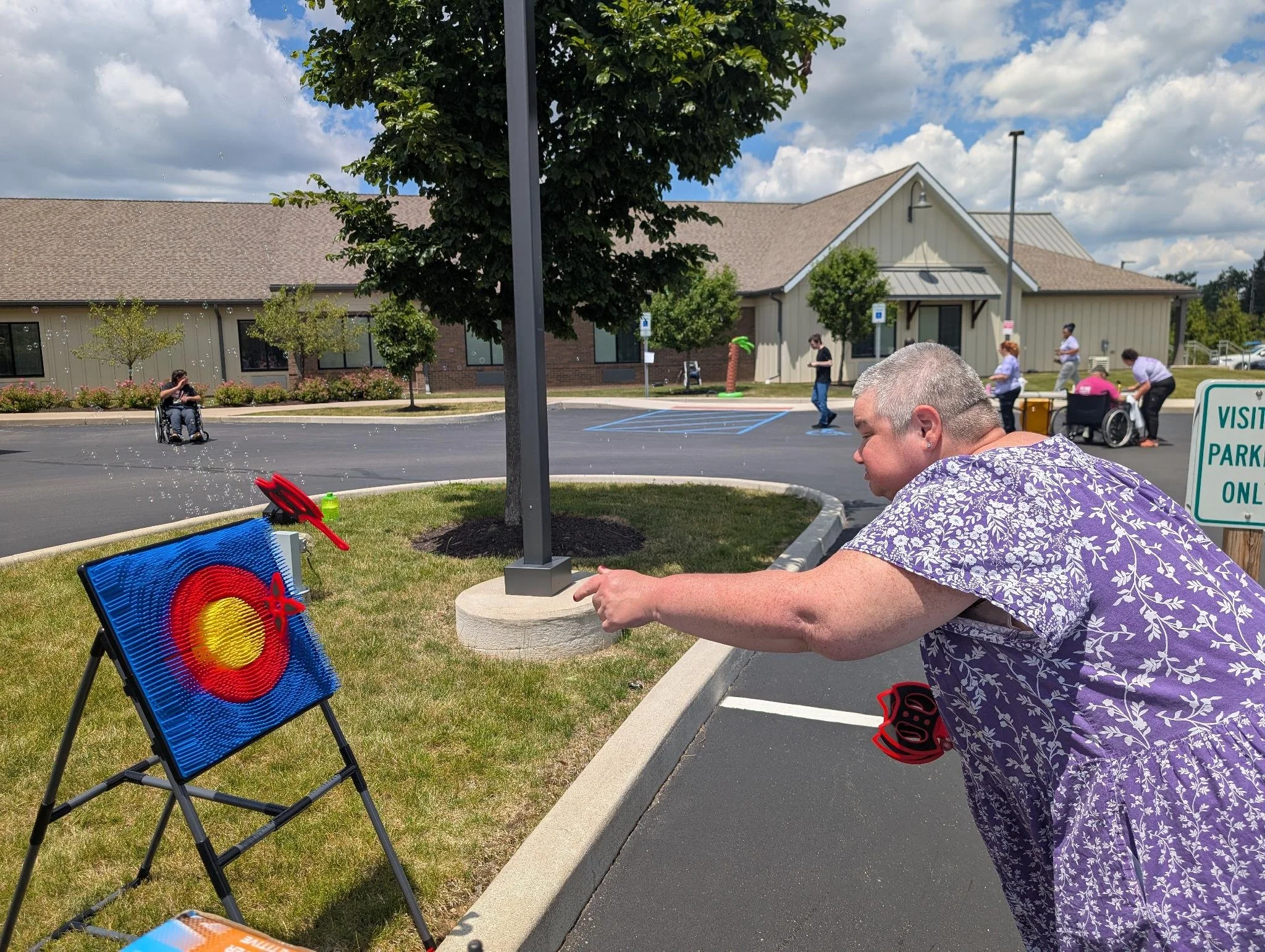 A woman with short gray hair and a purple patterned shirt is playing bean bag toss at an outdoor event. She is focused on aiming at a target on a stand. In the background, there are people walking and engaging in activities near a beige building, wit