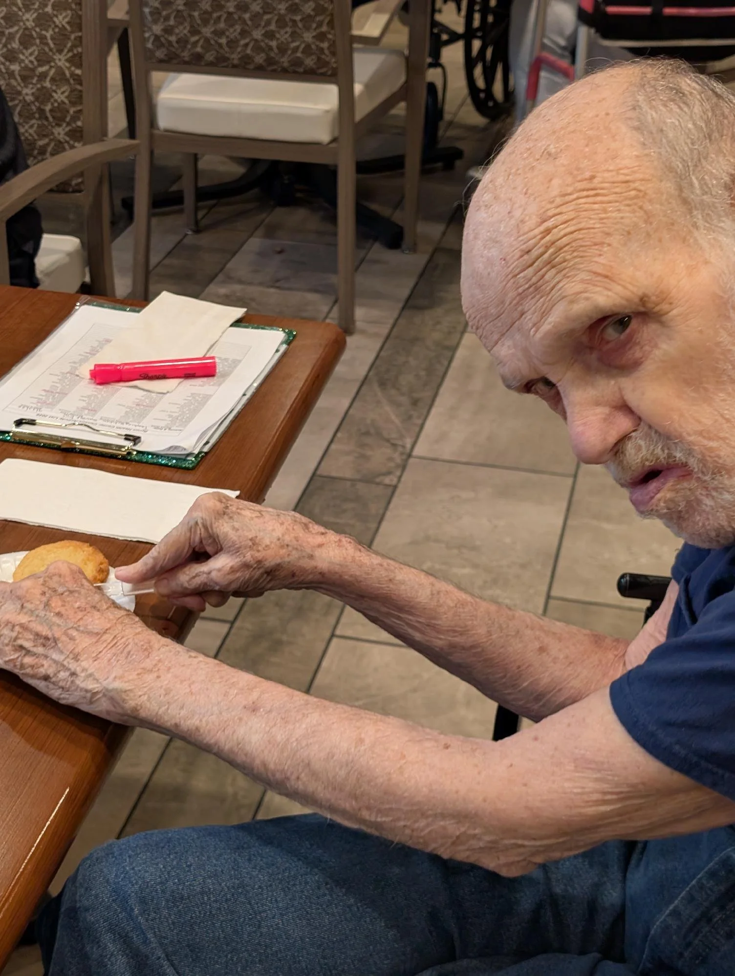 Close-up of elderly man's wrinkled hands and face as he points to a paper on a clipboard, sitting at a wooden table in a restaurant or cafe.