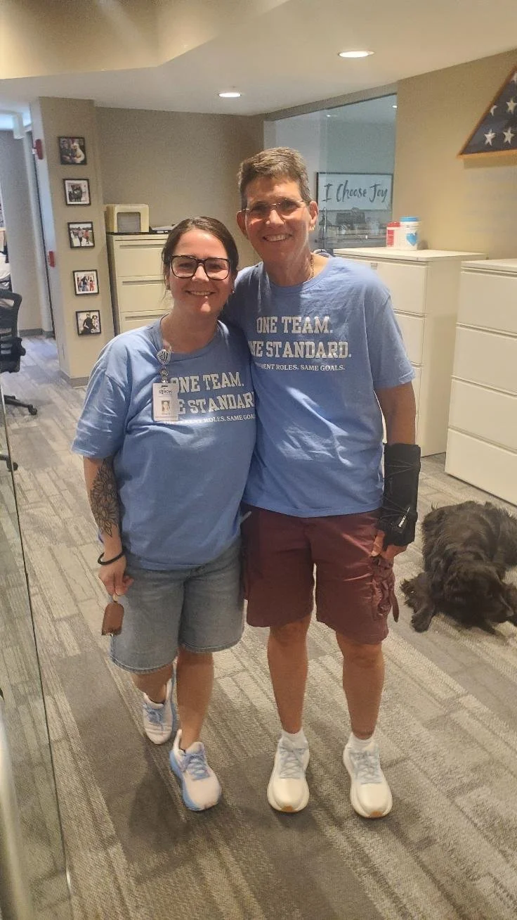 Two people standing side by side, smiling, in a room with filing cabinets and framed photos on the wall. Both are wearing matching blue T-shirts that say "ONE TEAM. ONE STANDARD." A dog is lying on the floor nearby.