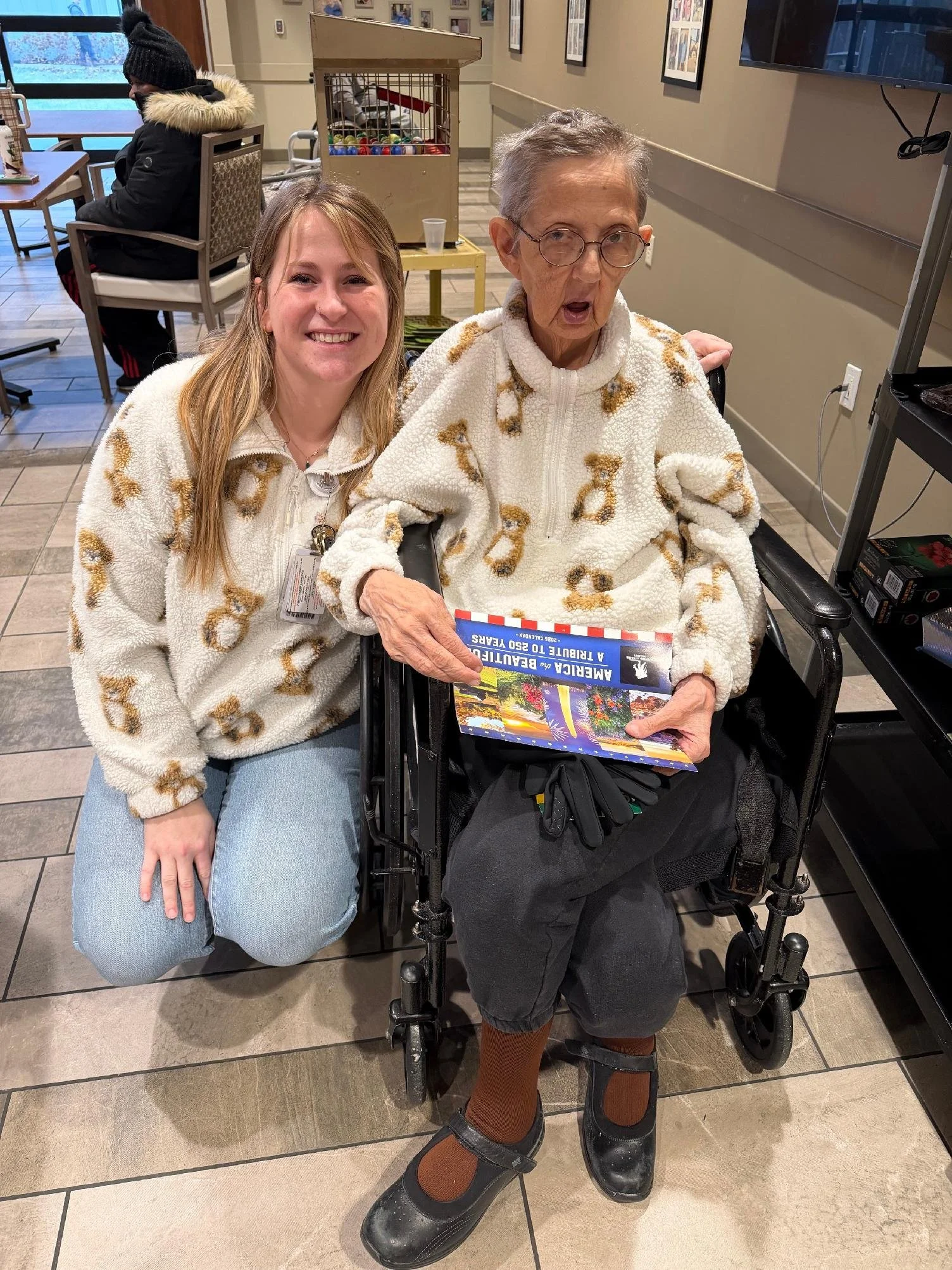 Two women, one young and one elderly, sitting together indoors. The elderly woman is in a wheelchair, holding a book about beach attractions, wearing a white fleece jacket with a leopard pattern, glasses, and black clogs. The young woman is kneeling beside her, smiling, and wearing a similar fleece jacket with a leopard pattern and light blue jeans. In the background, there is a woman sitting alone at a table near a window, with a gumball machine behind her.