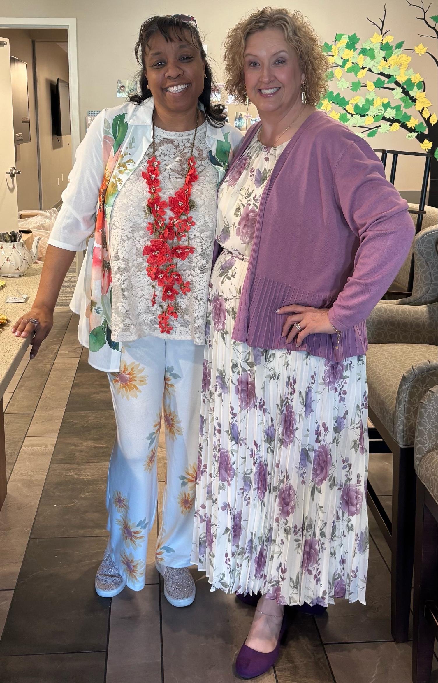 Two women smiling and posing together indoors, one wearing a floral embroidered suit and the other wearing a floral dress with a purple cardigan, standing in front of a wall with a tree and leaves artwork.