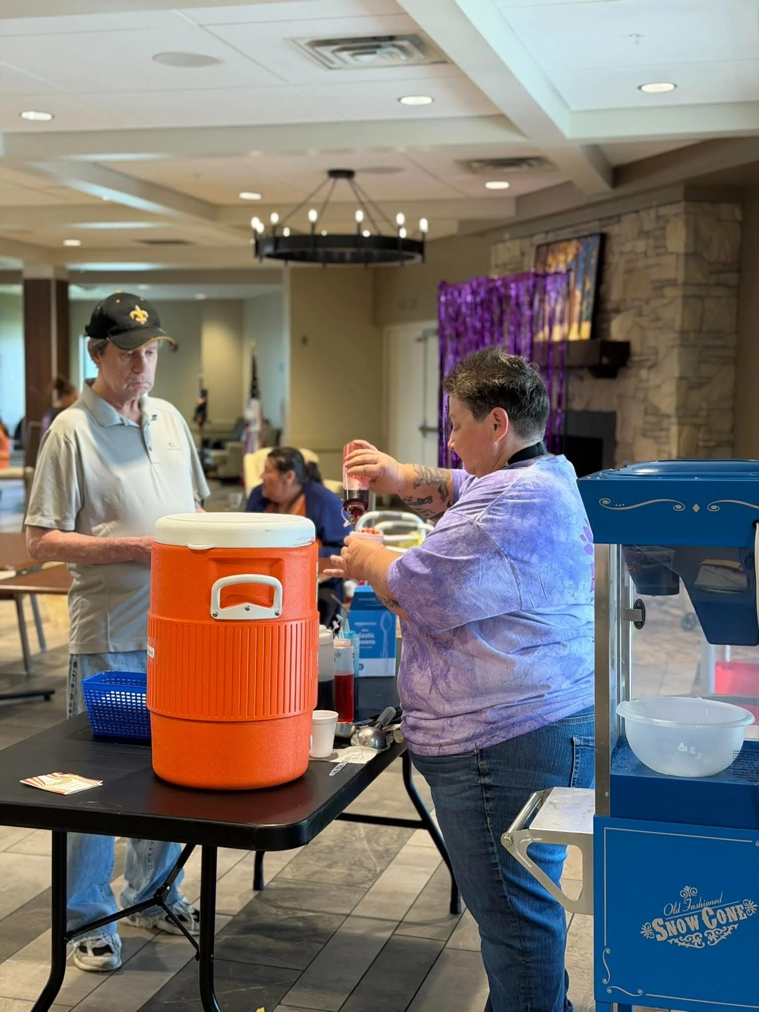 A man with tattoos on his arms and wearing a purple shirt is serving drinks at a snow cone stand, while another man with gray hair wearing a beige polo shirt and a black cap looks on. The stand has a blue sign with white lettering that says "Old Fash