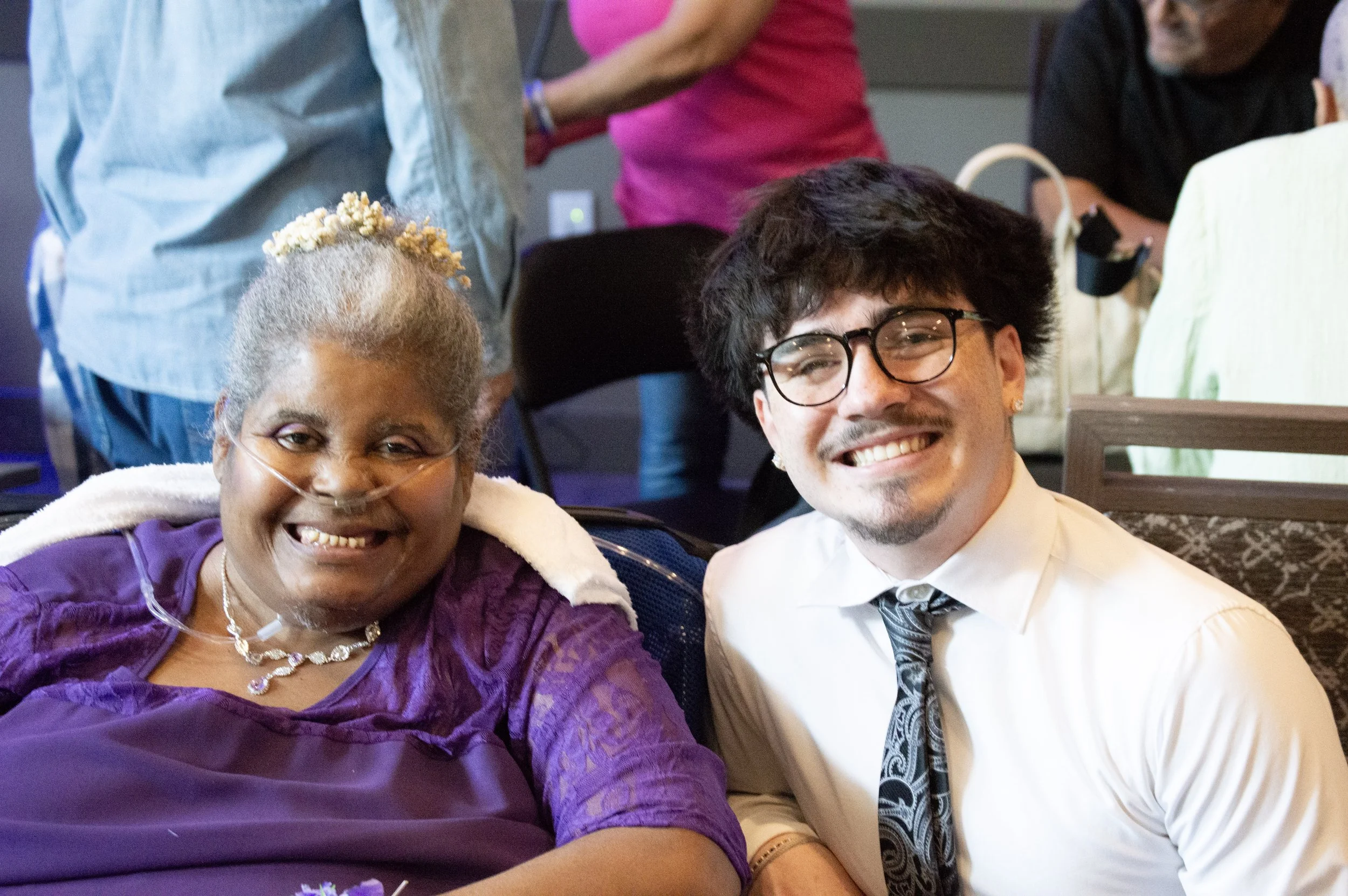 A joyful elderly woman with a nasal oxygen tube, smiling and wearing a purple dress with a necklace, sitting beside a young man with glasses, a white shirt, and a patterned tie, both taking a selfie at a social event.
