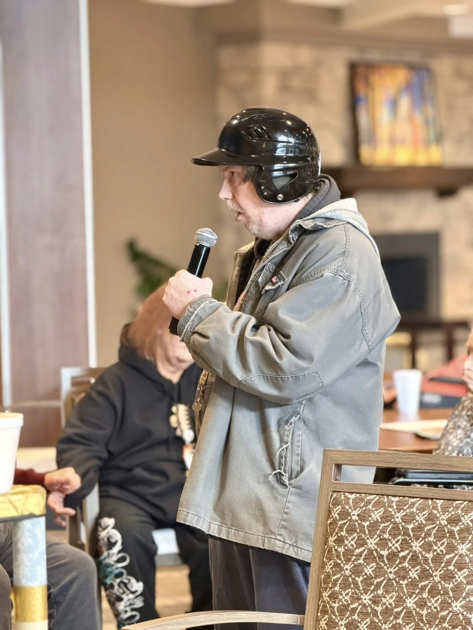 A man wearing a black safety helmet and gray jacket is speaking into a microphone in a restaurant or cafe setting. Several seated individuals are visible in the background.