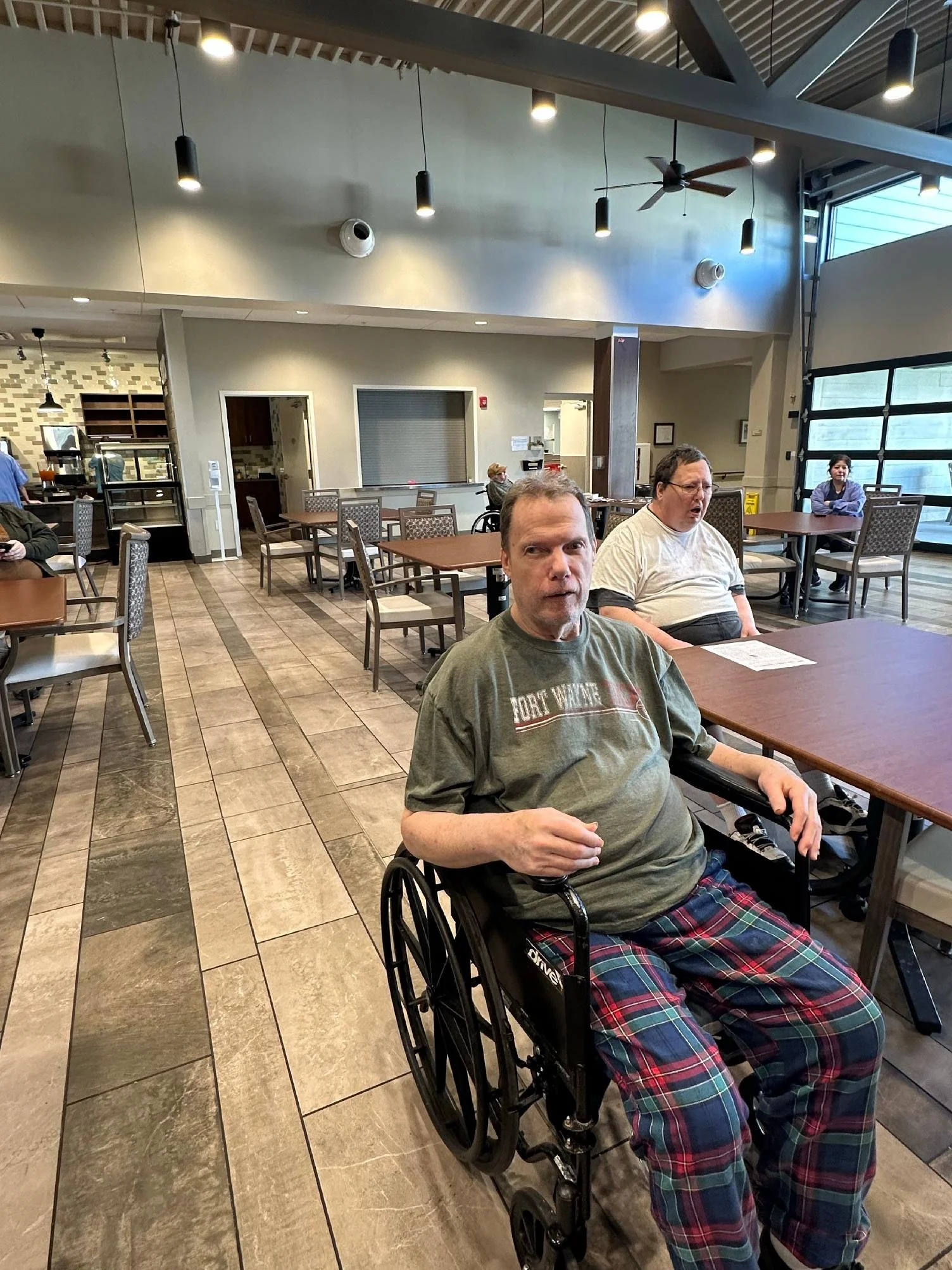 A man in a wheelchair wearing a gray t-shirt and plaid pajama pants sitting in a restaurant or cafe. Other people are seated at tables in the background, and the room has high ceilings with hanging lights and a ceiling fan.
