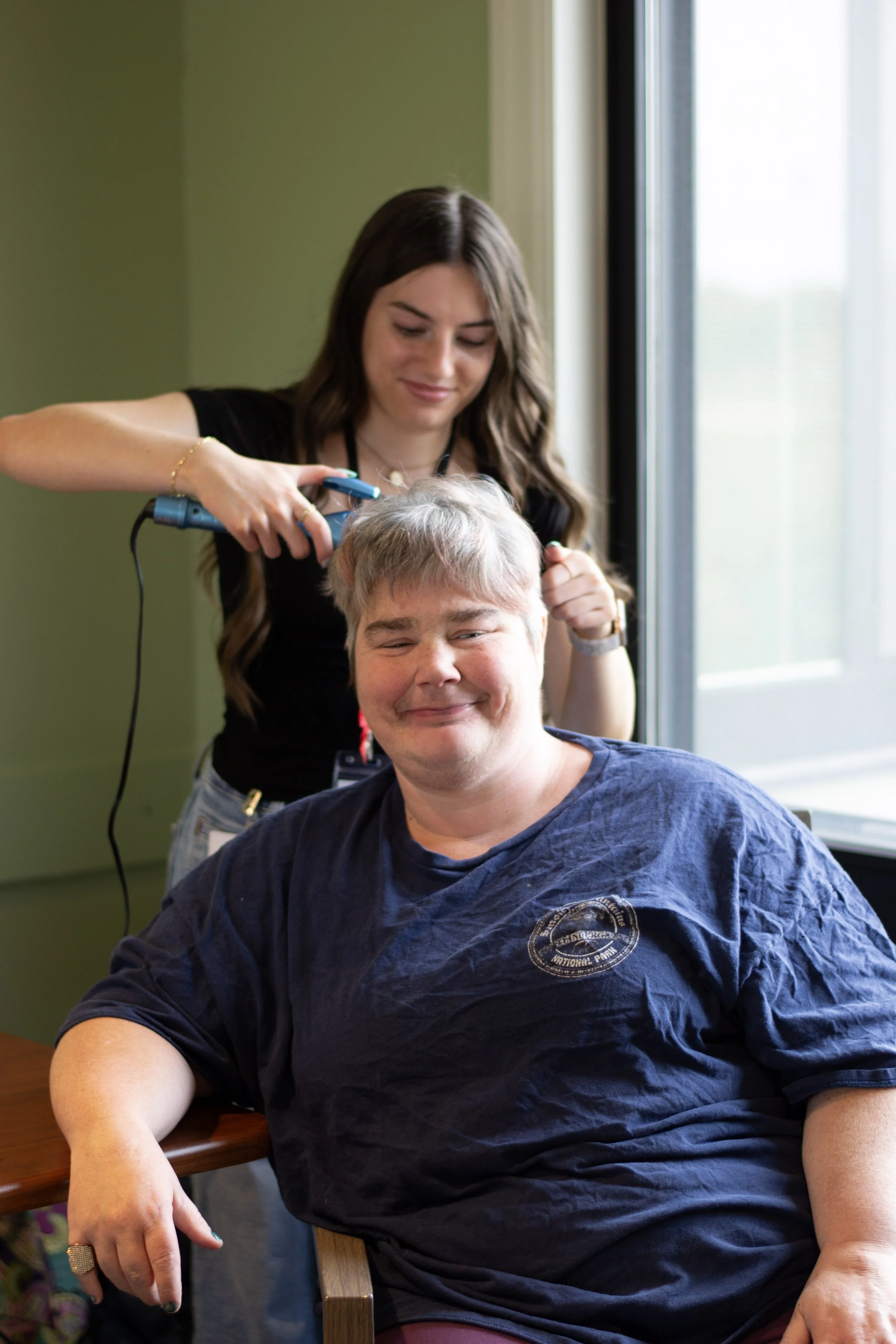 A young woman with long brown hair is giving a haircut to a man with graying hair, sitting at a table near a window, with a green wall in the background.