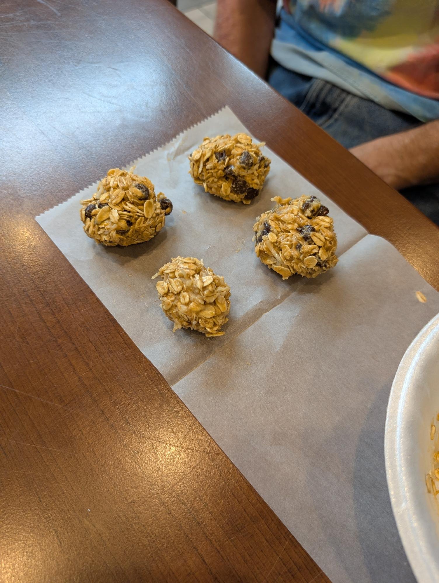 Four oatmeal cookie energy bites with chocolate chips on a piece of parchment paper on a wooden table, with a person wearing a tie-dye shirt visible in the background.