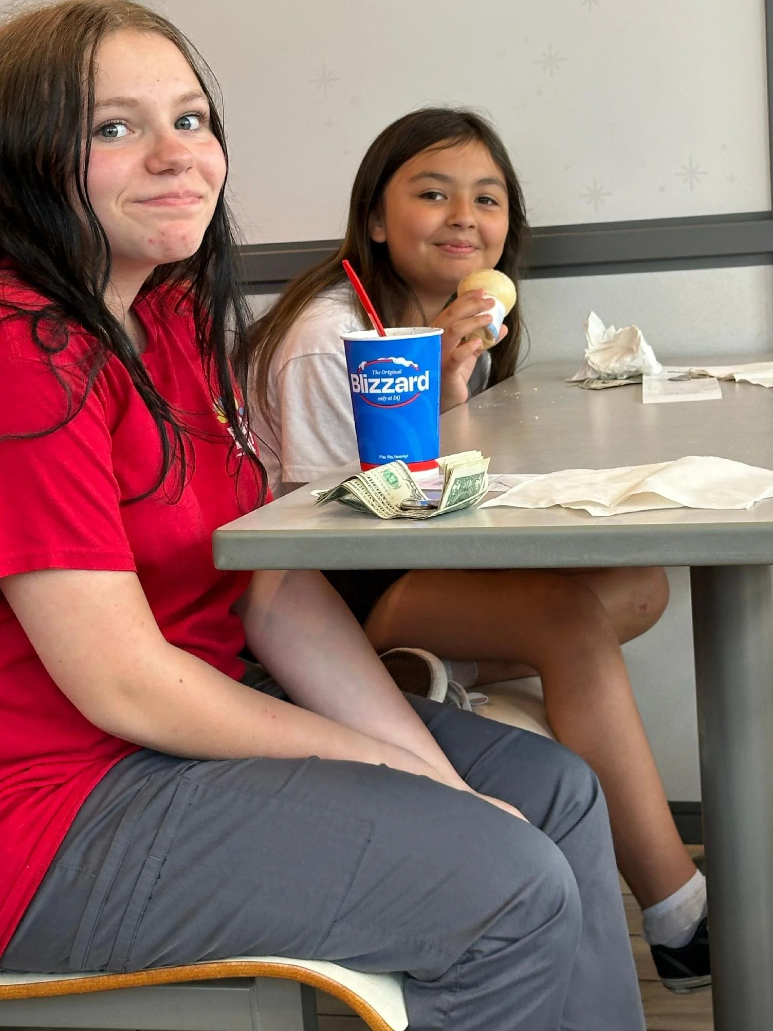 Two young girls sitting at a table, one holding an ice cream cone and the other with a cup of soft serve ice cream, with money and crumpled napkins on the table.