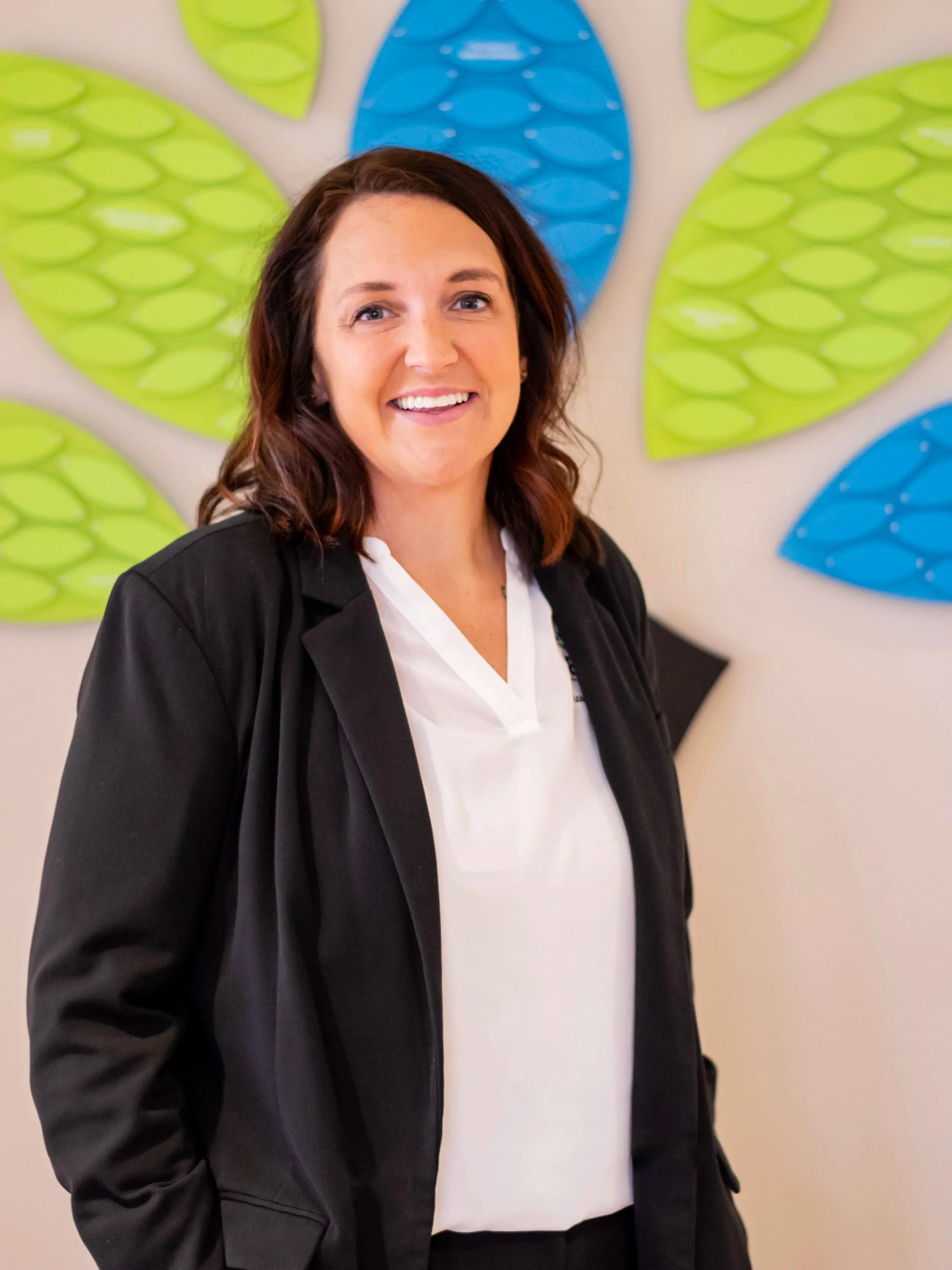 A woman with shoulder-length brown hair, wearing a black blazer over a white blouse, is smiling at the camera. Behind her is a wall with an abstract art piece featuring large, leaf-like shapes in green and blue colors.