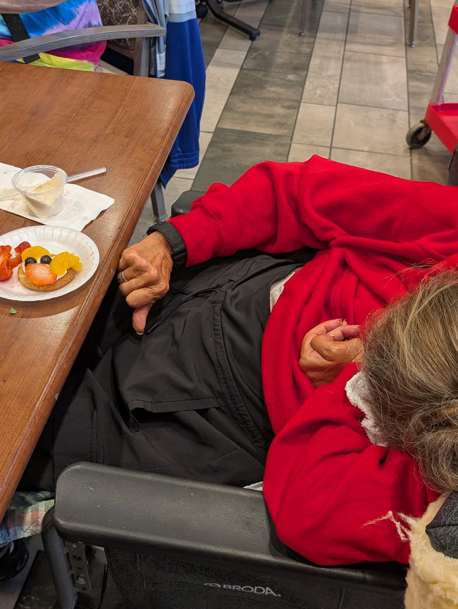 A person dressed in a red jacket and Santa hat sitting in a wheelchair at a table in a restaurant or café. Their right hand is resting on their lap, and the person's head is turned away from the camera. On the table, there's a plate with a fruit-topp