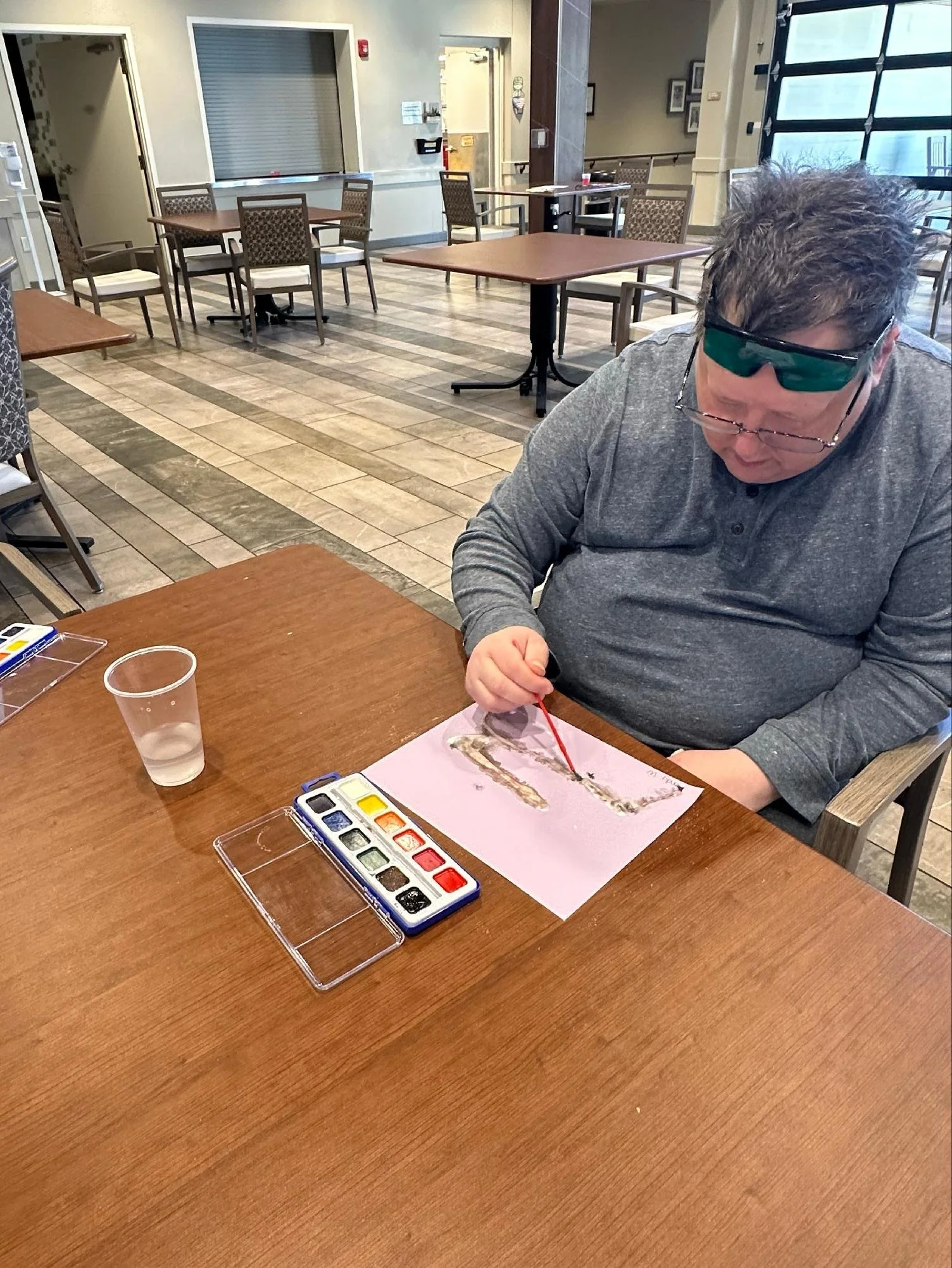 A man wearing dark sunglasses and a gray long-sleeve shirt sits at a wooden table, painting with watercolor paints on a pink sheet of paper. A plastic cup of water is on the table, and a watercolor paint set is open nearby. The room has multiple empt