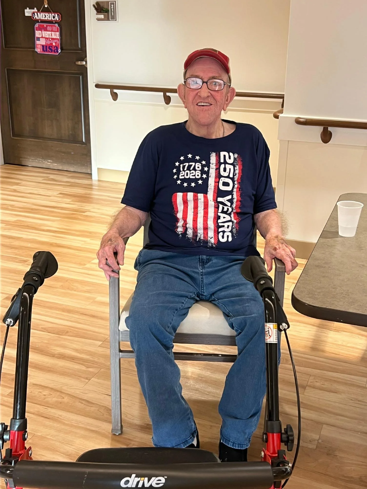 An elderly man with glasses, a red cap, and a blue t-shirt with American flags and the words '1776 2026 250 YEARS' is sitting in a wheelchair inside a room. The room has hardwood floors, a wall with a small hanging shelf with decorative items, and pa