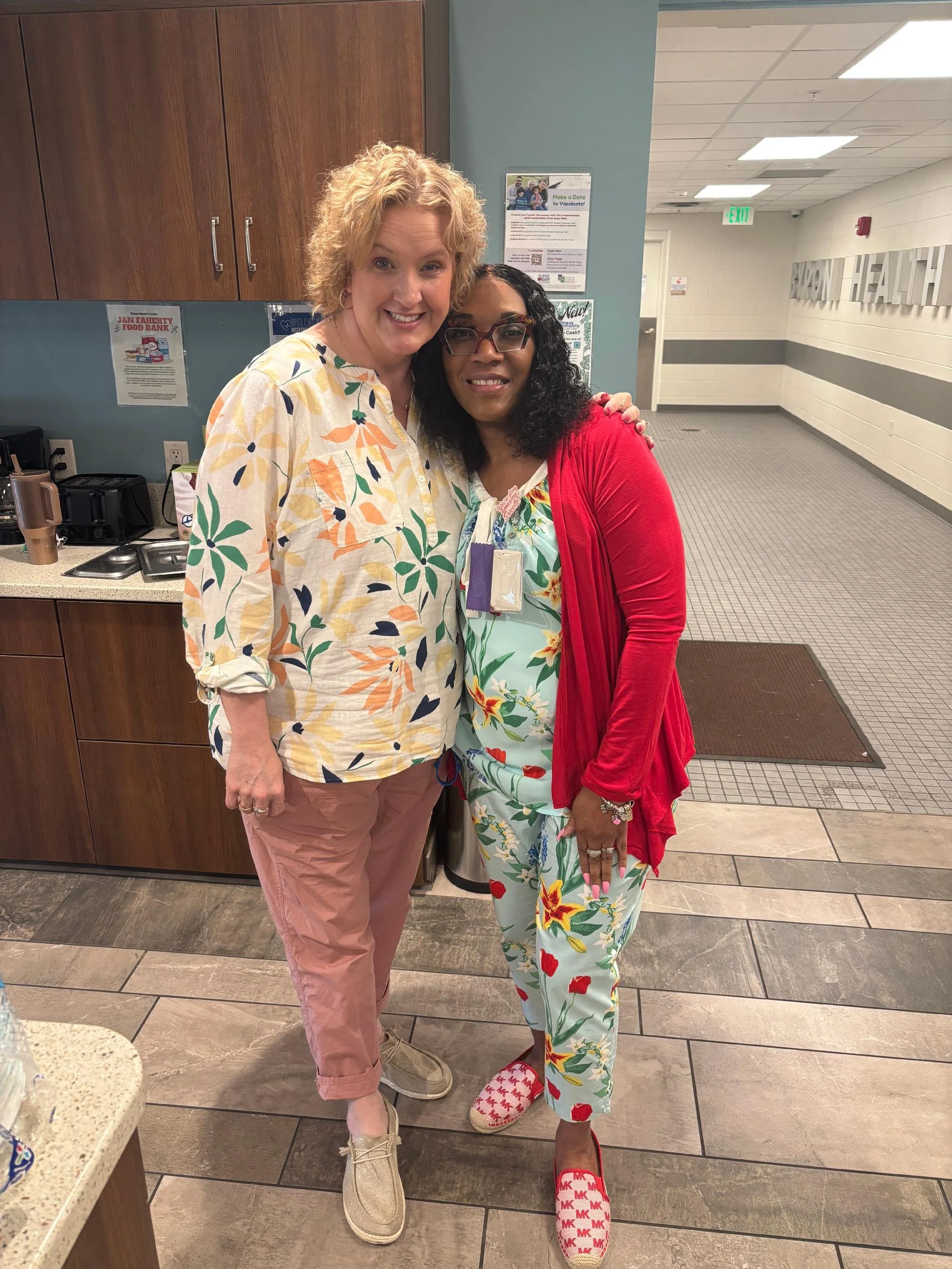Two women standing inside a building, smiling at the camera. The woman on the left has curly blonde hair and is wearing a light-colored floral shirt with pink pants. The woman on the right has curly black hair, glasses, and is dressed in colorful flo