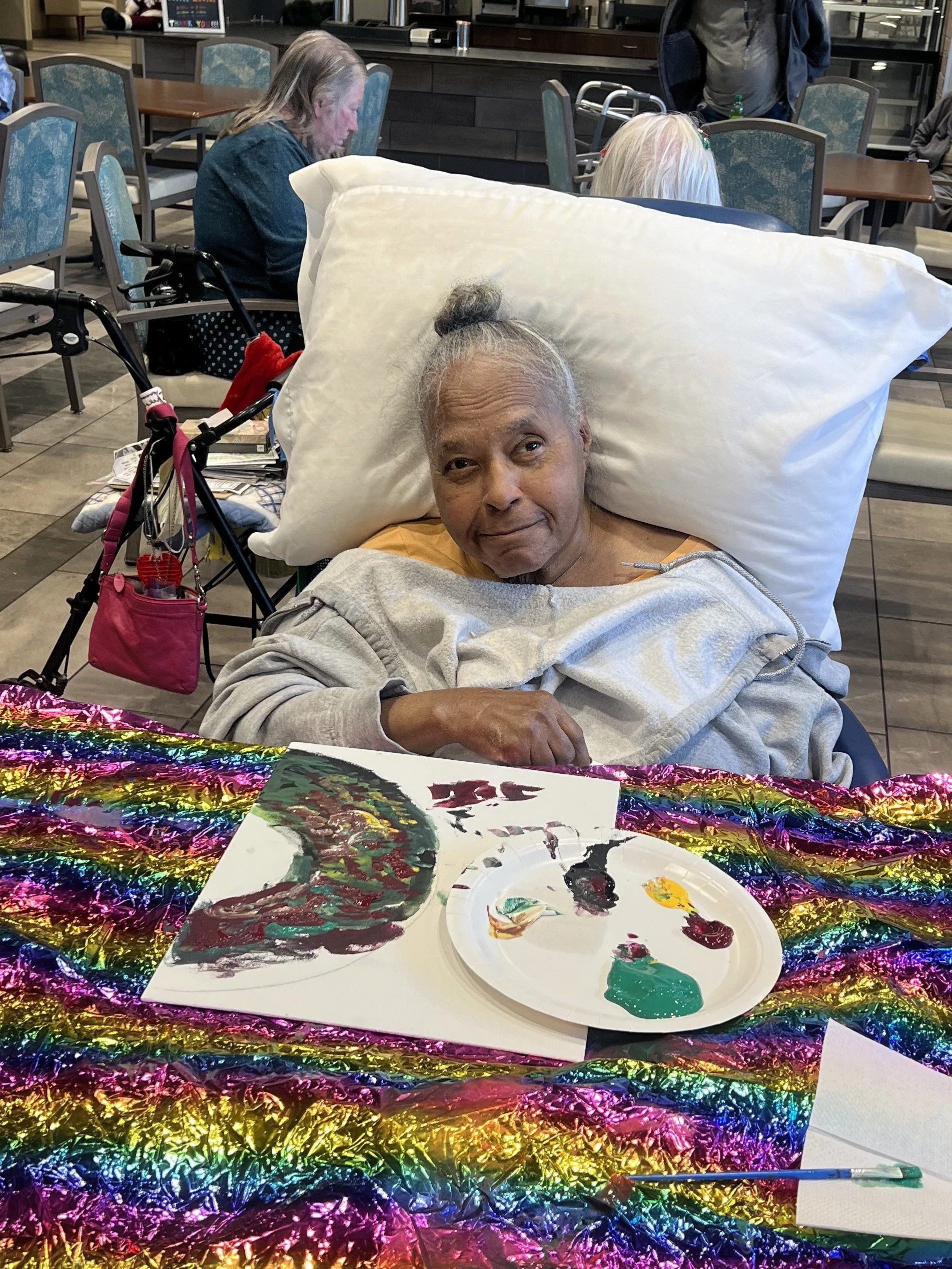 Elderly woman sitting at a table with arts and crafts supplies, including a paper plate with paint and a paintbrush. She has a large pillow on her head and is smiling. The table is decorated with shiny rainbow-colored material, and in the background, other people are sitting in a communal space with tables and chairs.