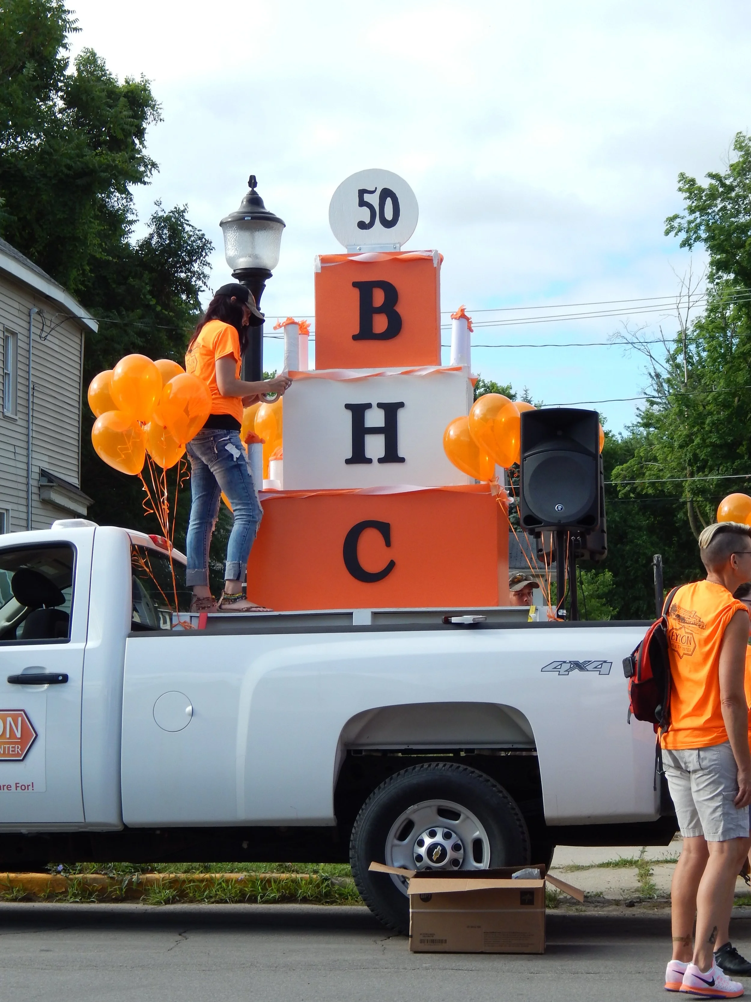 A parade float on a white pickup truck decorated in orangesurrounded by orange balloons. A person in an orange shirt and black cap is standing on the float, adjusting something. Large cardboard boxes are on the ground in front of the truck, with a woman in white shorts and a bright orange vest standing nearby.