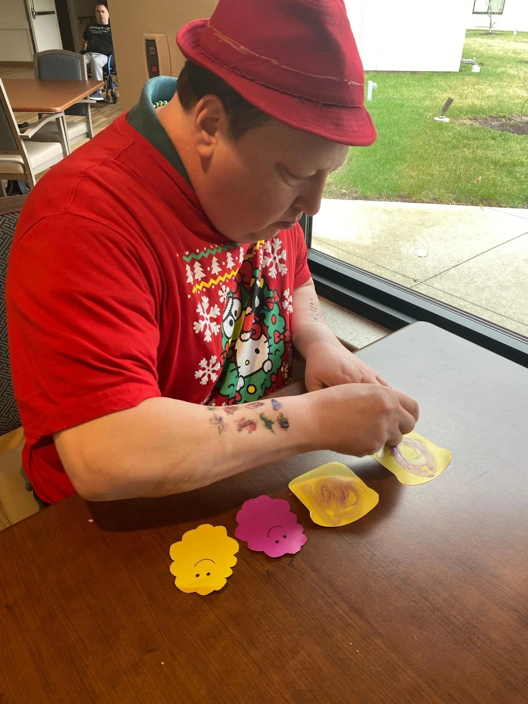A person wearing a red hat and a Christmas-themed shirt is sitting at a table, decorating paper flowers with colorful swirls on them. There are additional paper flowers on the table, and a large window shows a green lawn outside.