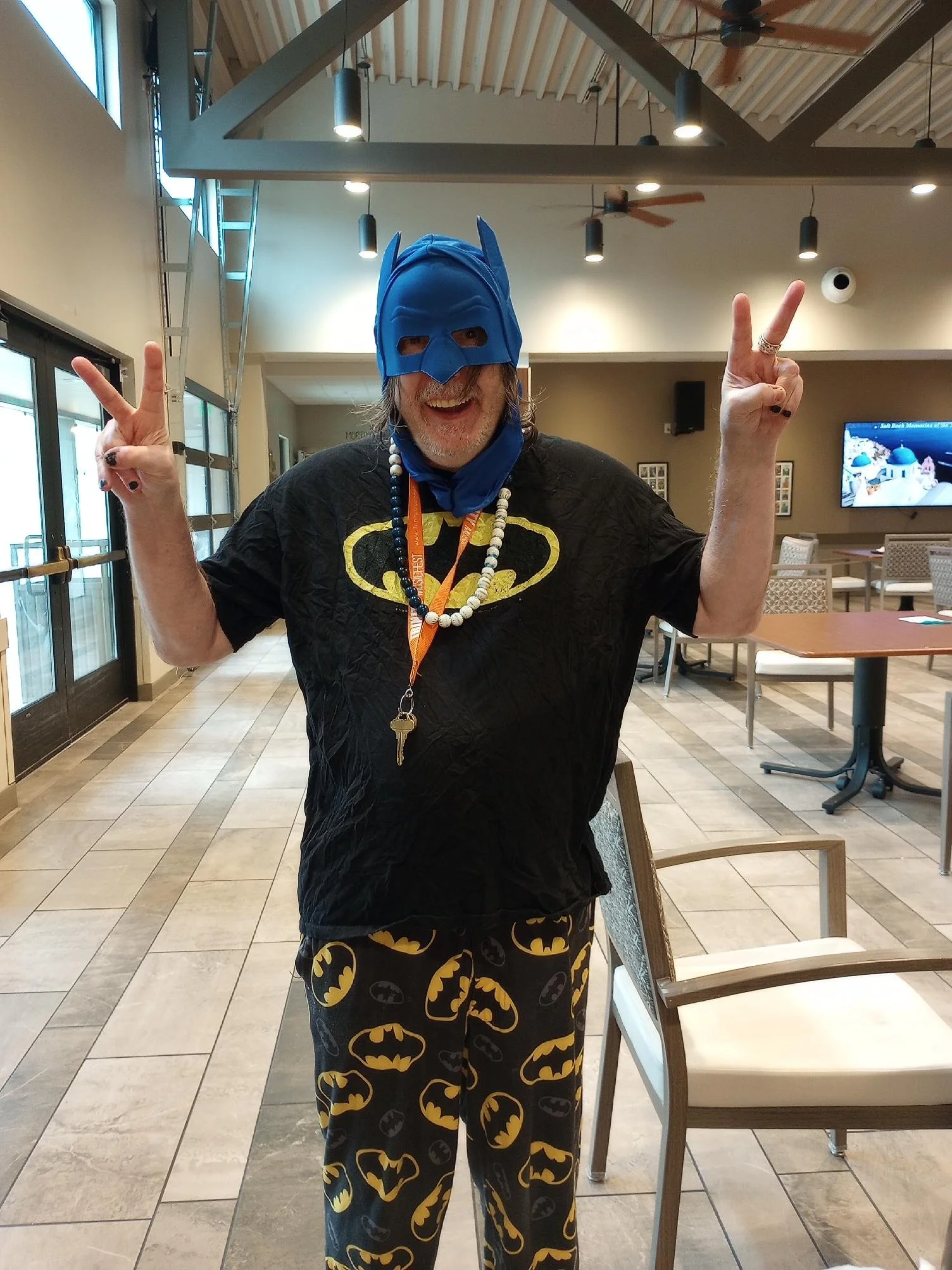 Person wearing a Batman mask, making peace signs with both hands, dressed in Batman-themed clothing, inside a restaurant or cafe.