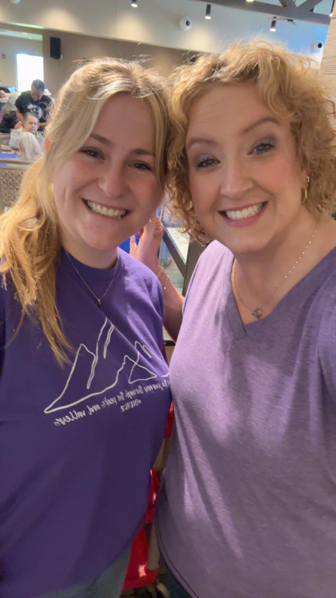Close-up selfie of two smiling women in purple shirts with a group of people in the background at an indoor venue.