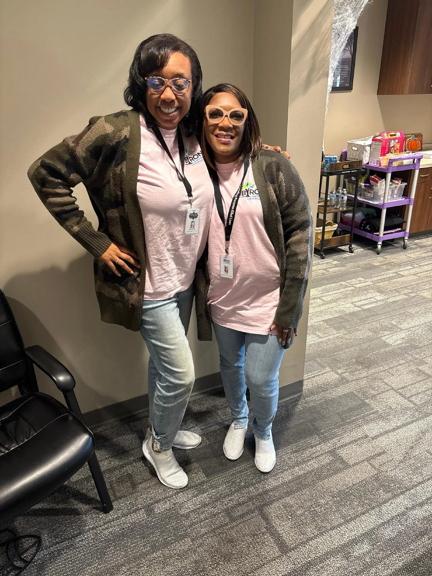 Two women standing indoors in front of a beige wall, smiling and posing. They are both wearing pink shirts with a logo, glasses, and gray sweaters or jackets. The woman on the left has short black hair, while the woman on the right has shoulder-length hair. There are chairs and a cart with supplies in the background.
