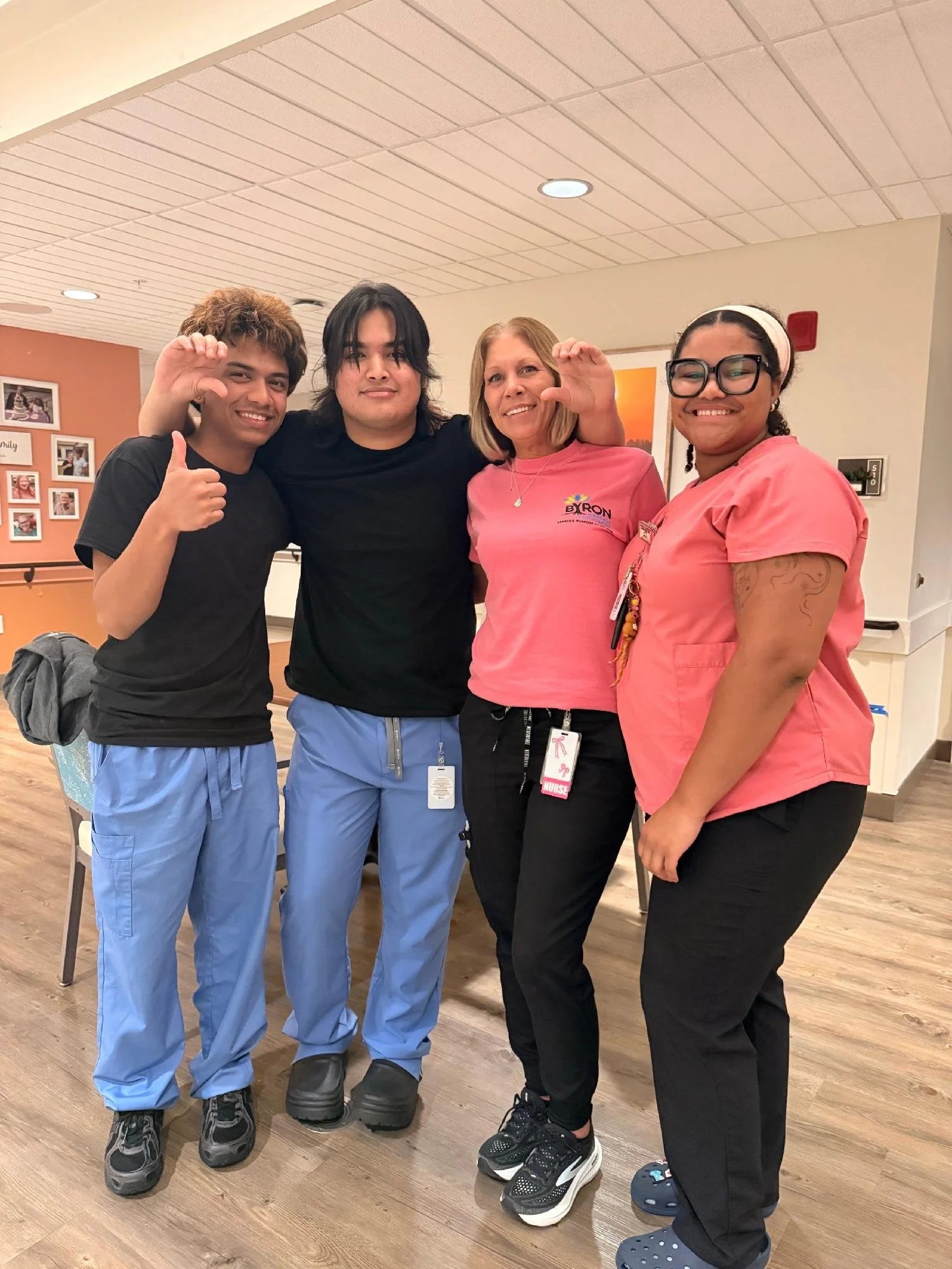 Four people, including two women wearing medical scrubs, posing indoors, smiling, and making a waving gesture.