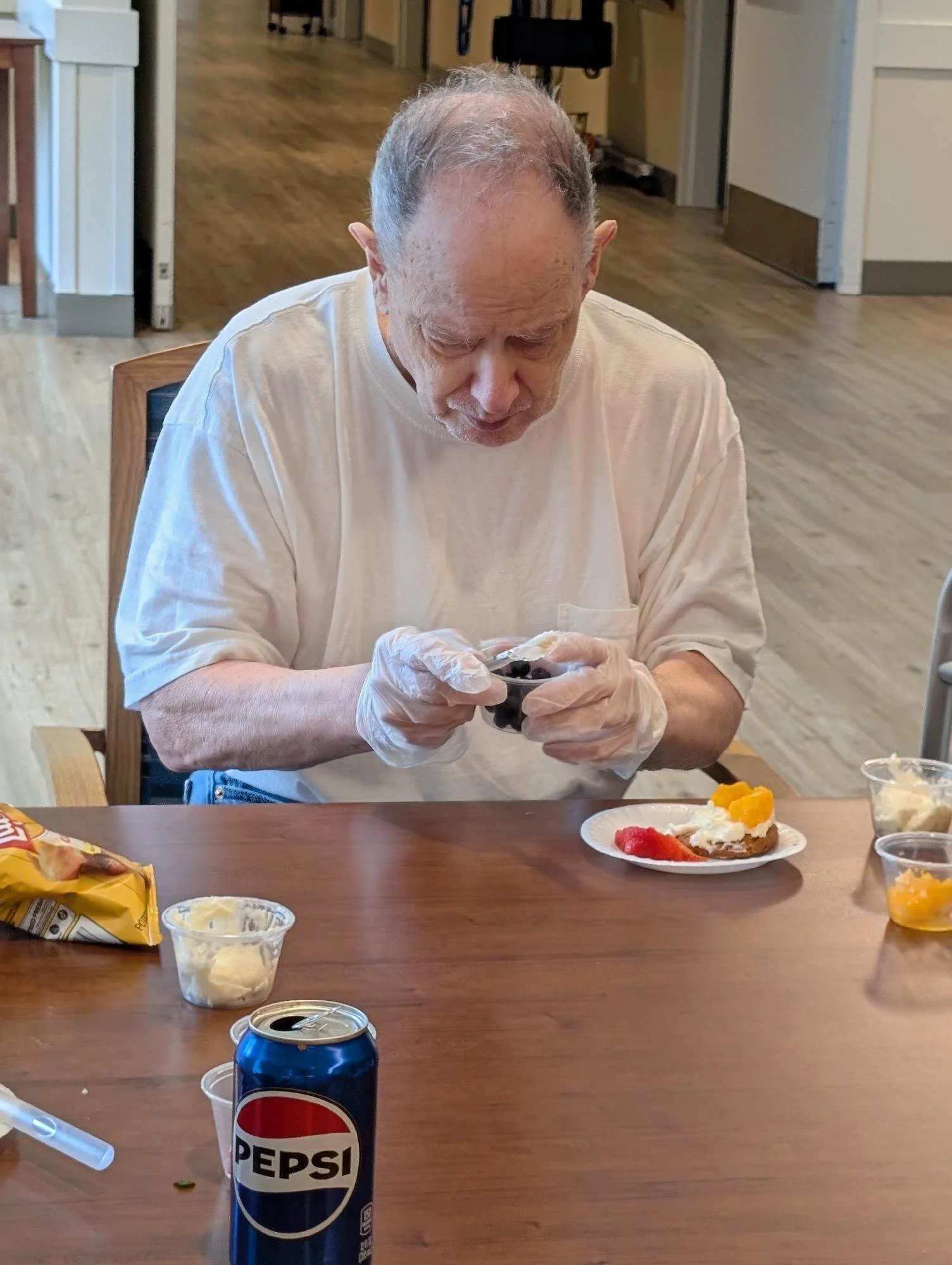 An elderly man with gray hair, wearing a white shirt and gloves, is sitting at a table and looking at a small bowl of food. There is a plate with a dessert topped with whipped cream, strawberries, and peaches in front of him. On the table, there are 