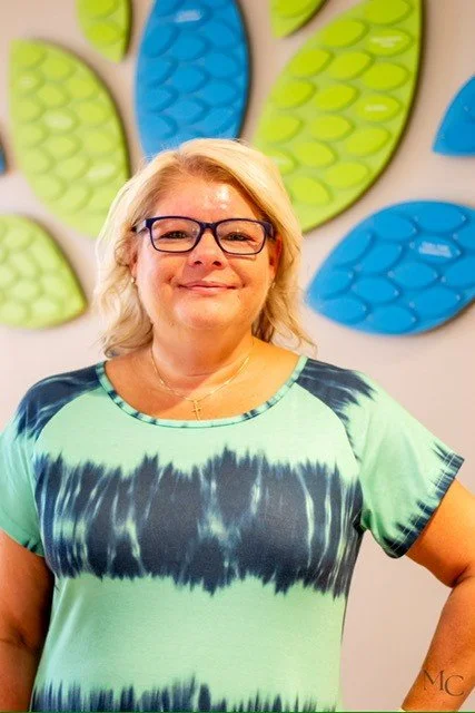 A woman with glasses and shoulder-length blonde hair standing in front of a brightly colored wall art with abstract shapes in green and blue, wearing a tie-dye style green and black shirt.