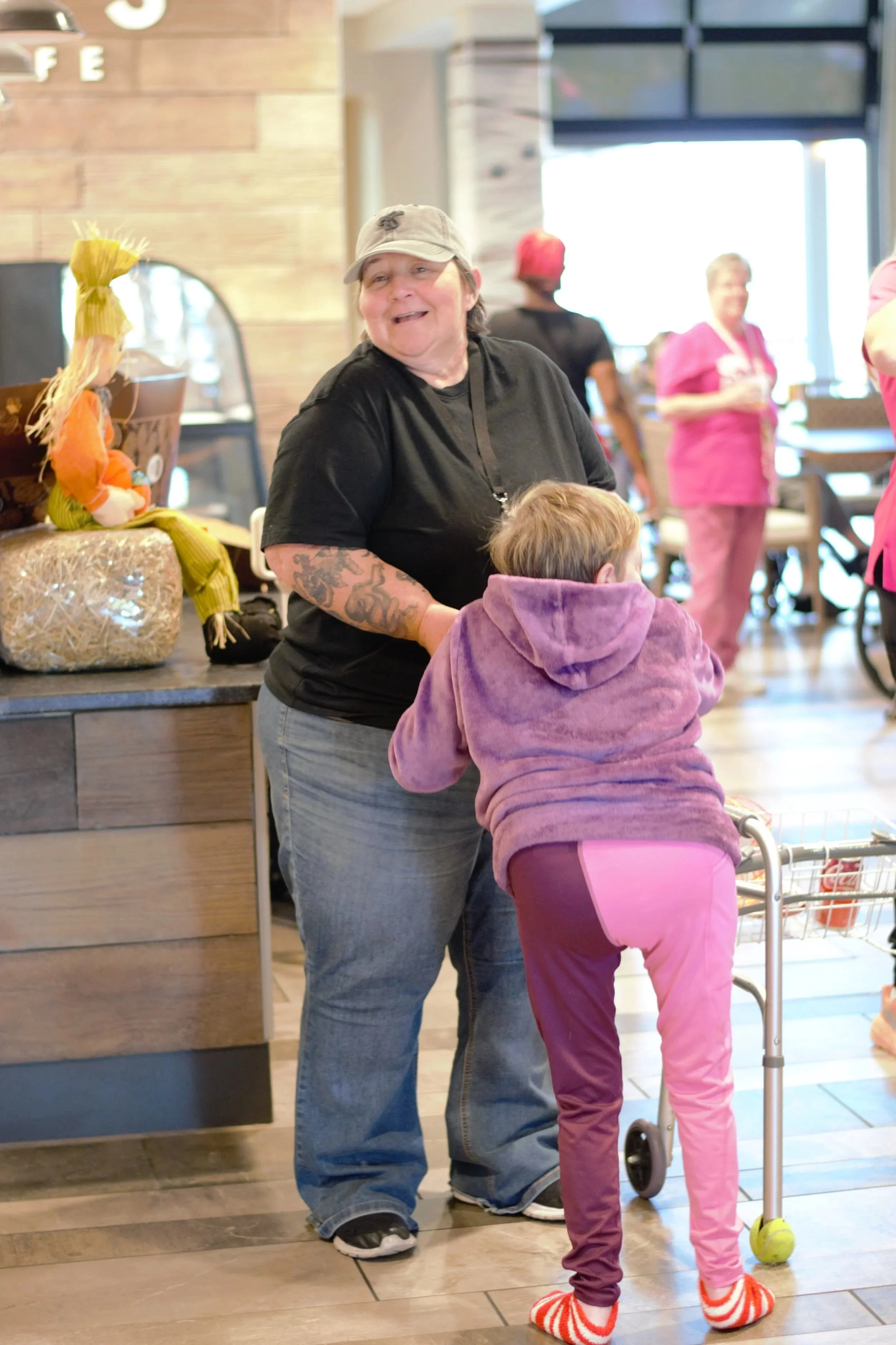 A woman in a gray baseball cap and black t-shirt smiling while a young girl with short hair in a purple hoodie stands with her back to the camera. The girl is using a walker and wearing striped socks. The scene is inside a coffee shop or restaurant with blurred patrons in the background.
