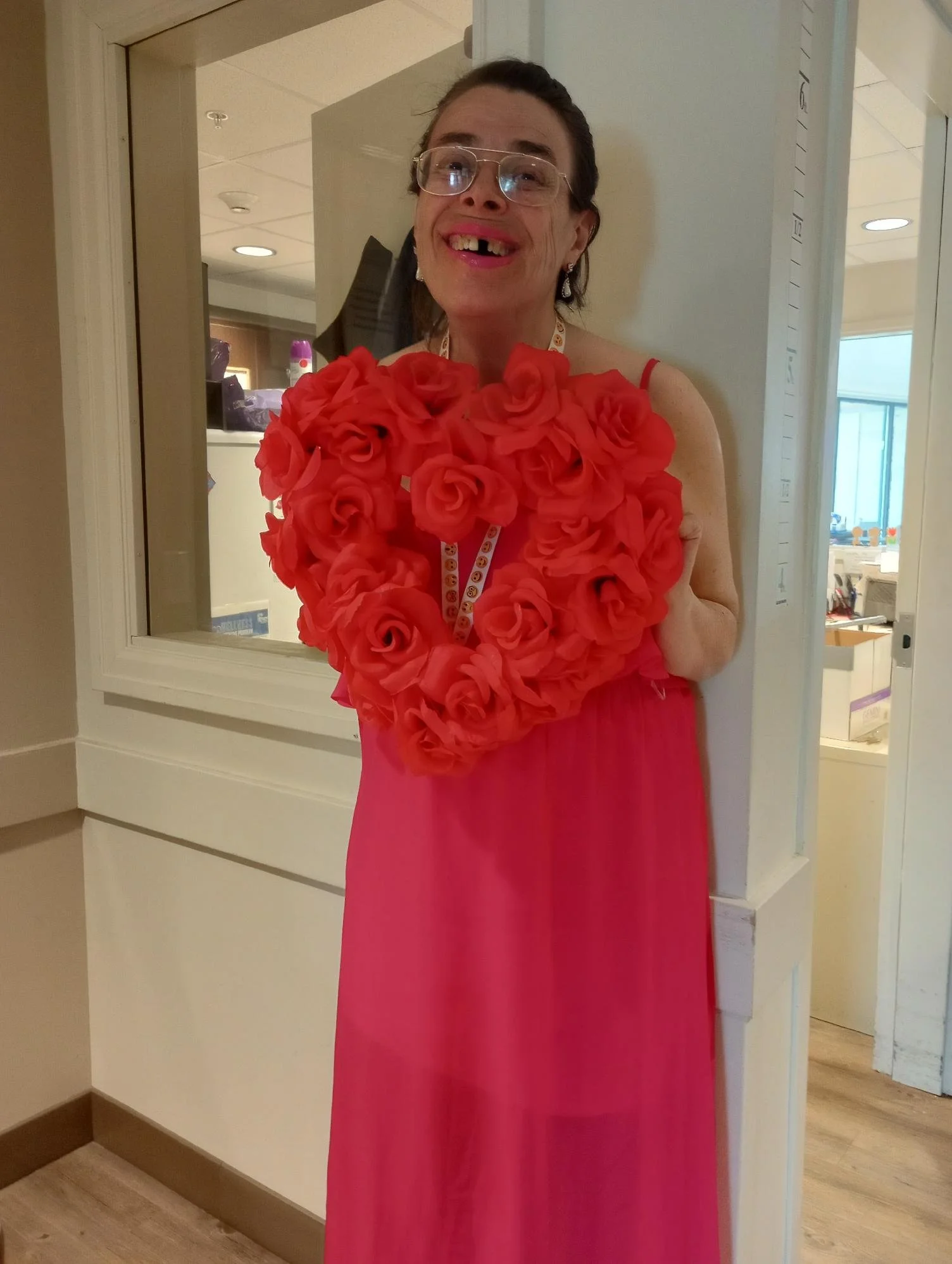 An elderly woman with glasses, missing two front teeth, dressed in a pink dress with an orange rose flower collar, is smiling through a square window or opening in a wall.