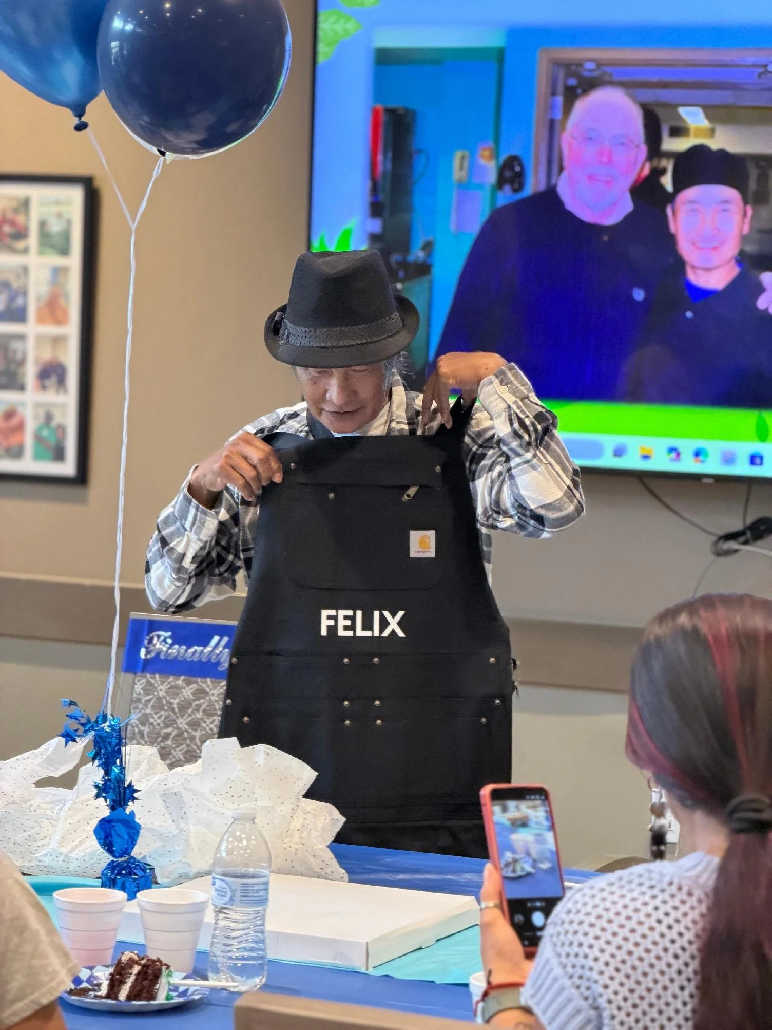 A person wearing a black hat and plaid shirt opening a gift bag with the name 'FELIX' on it at a celebration table decorated with balloons and cake. In the background, a large monitor shows a photo of two people smiling, with one person wearing glass