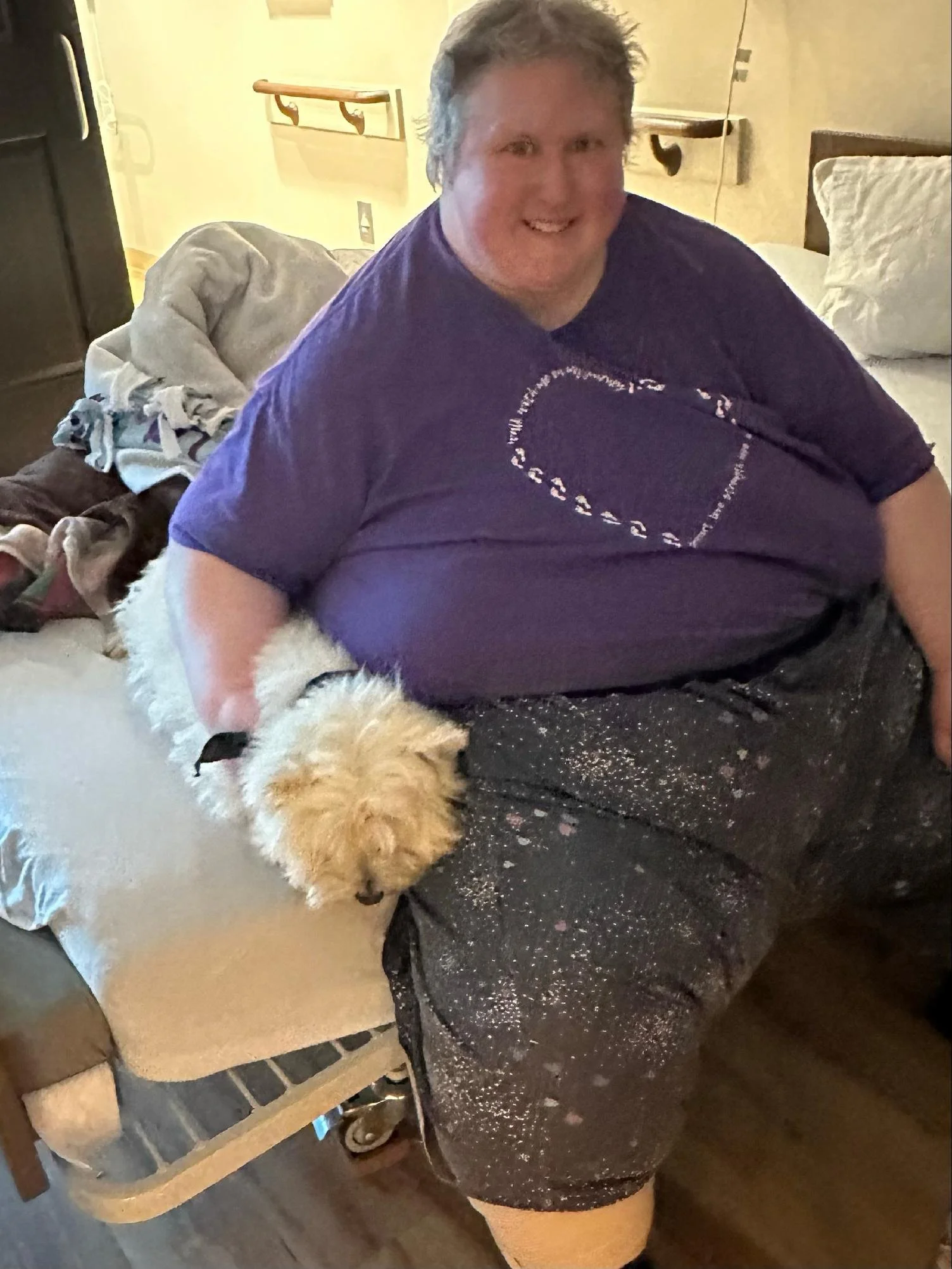 A person with short hair smiling, wearing a purple t-shirt, sitting on the floor with a small fluffy dog near their leg, in a room with a bed, pillow, and a blanket in the background.