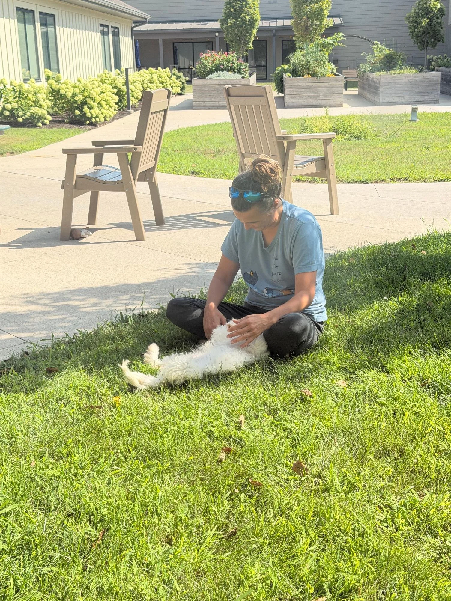 A woman sitting on the grass petting a white dog with black spots. Two empty wooden chairs are positioned on a paved patio in the background, with a house and some planters filled with flowers and greenery behind them.