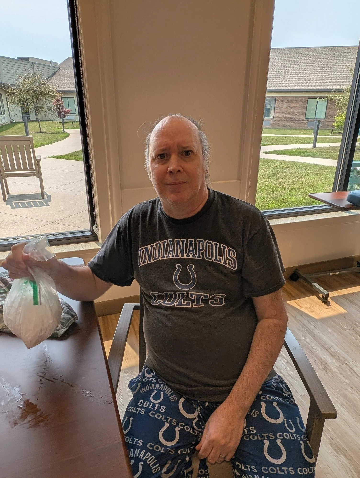 A middle-aged man with gray hair is sitting at a table inside a room, holding a plastic bag. He is wearing a dark gray T-shirt with 'Indianapolis Colts' written on it and matching shorts with the Colts logo. There is a large window behind him showing