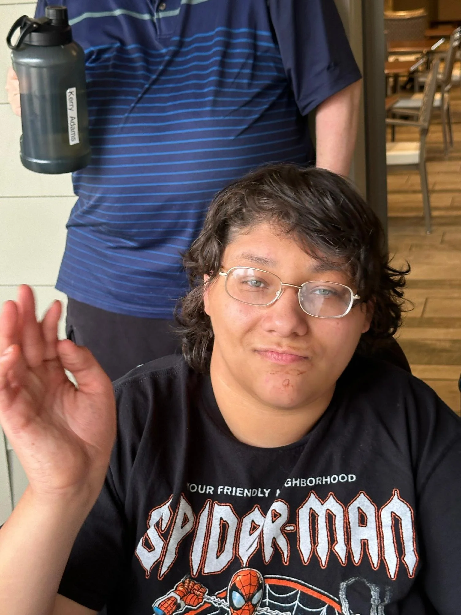 A young man with glasses and curly hair waving at the camera, wearing a black Spider-Man t-shirt with orange and white lettering and graphics, in an indoor setting with wooden flooring and chairs in the background.