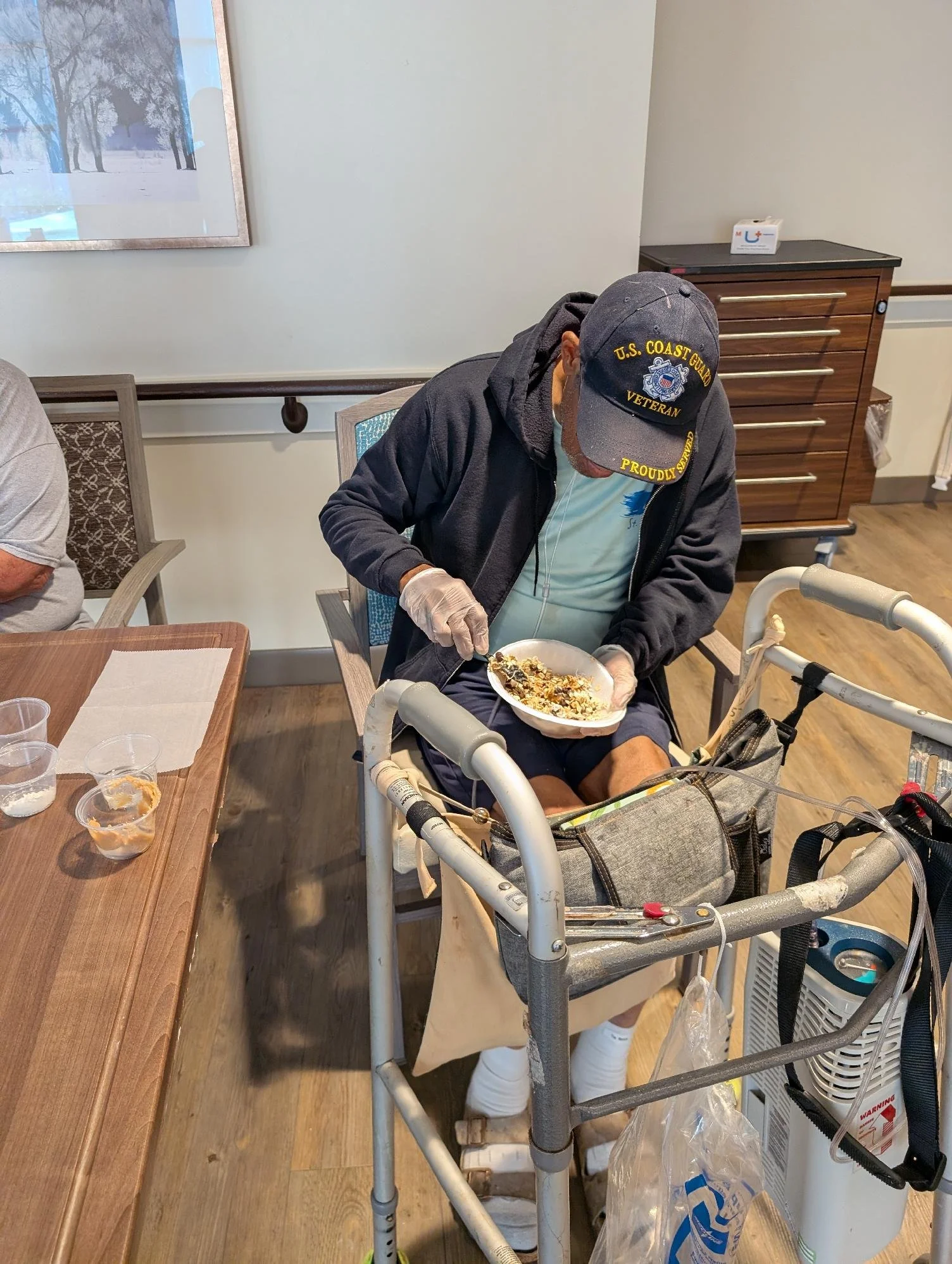 A person in a wheelchair wearing a U.S. Coast Guard veteran hat serving food with gloves in a dining area at a care facility.