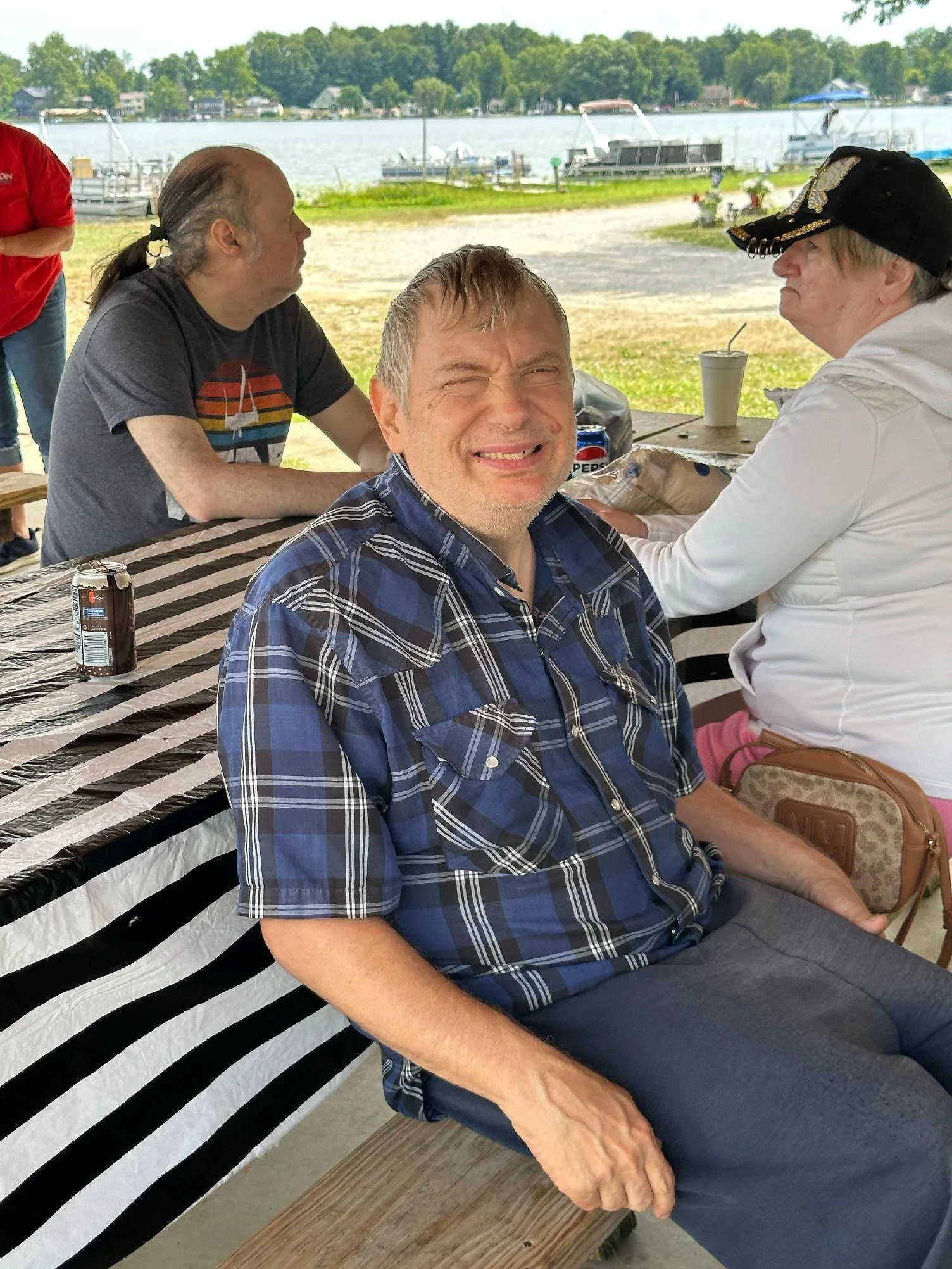 A group of people sitting outdoors by a lake, with boats on the water and trees in the background. One man in a plaid shirt is smiling at the camera, while others are engaged in conversation.