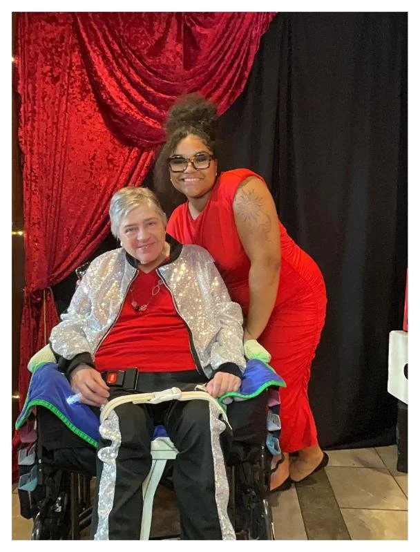 Two women, one in a wheelchair, smiling and posing for a photo in front of red and black curtains.