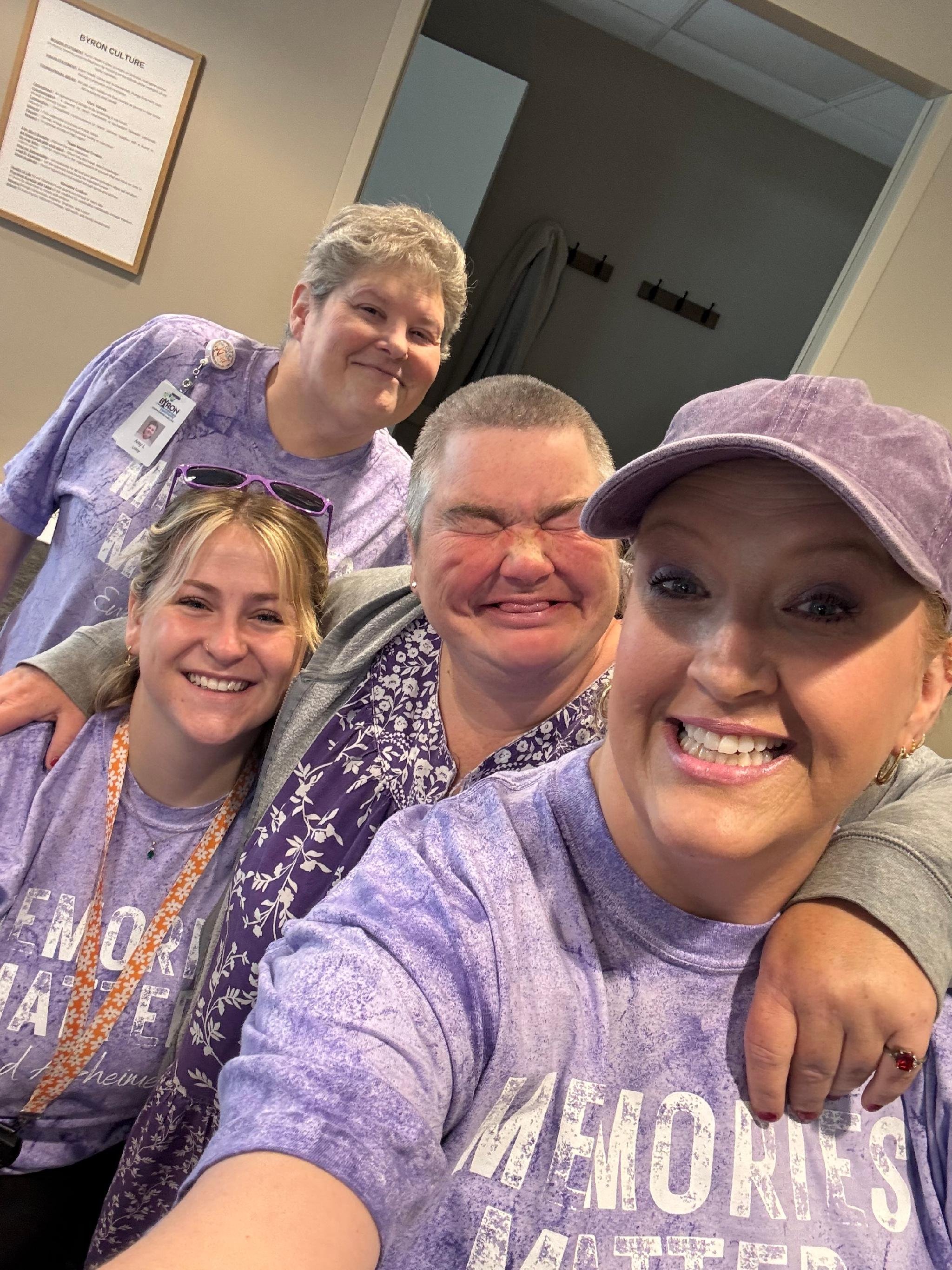 A group of four smiling women taking a selfie together indoors. One woman is wearing a purple cap, another has sunglasses on her head, and some are wearing purple shirts. They appear happy and friendly.