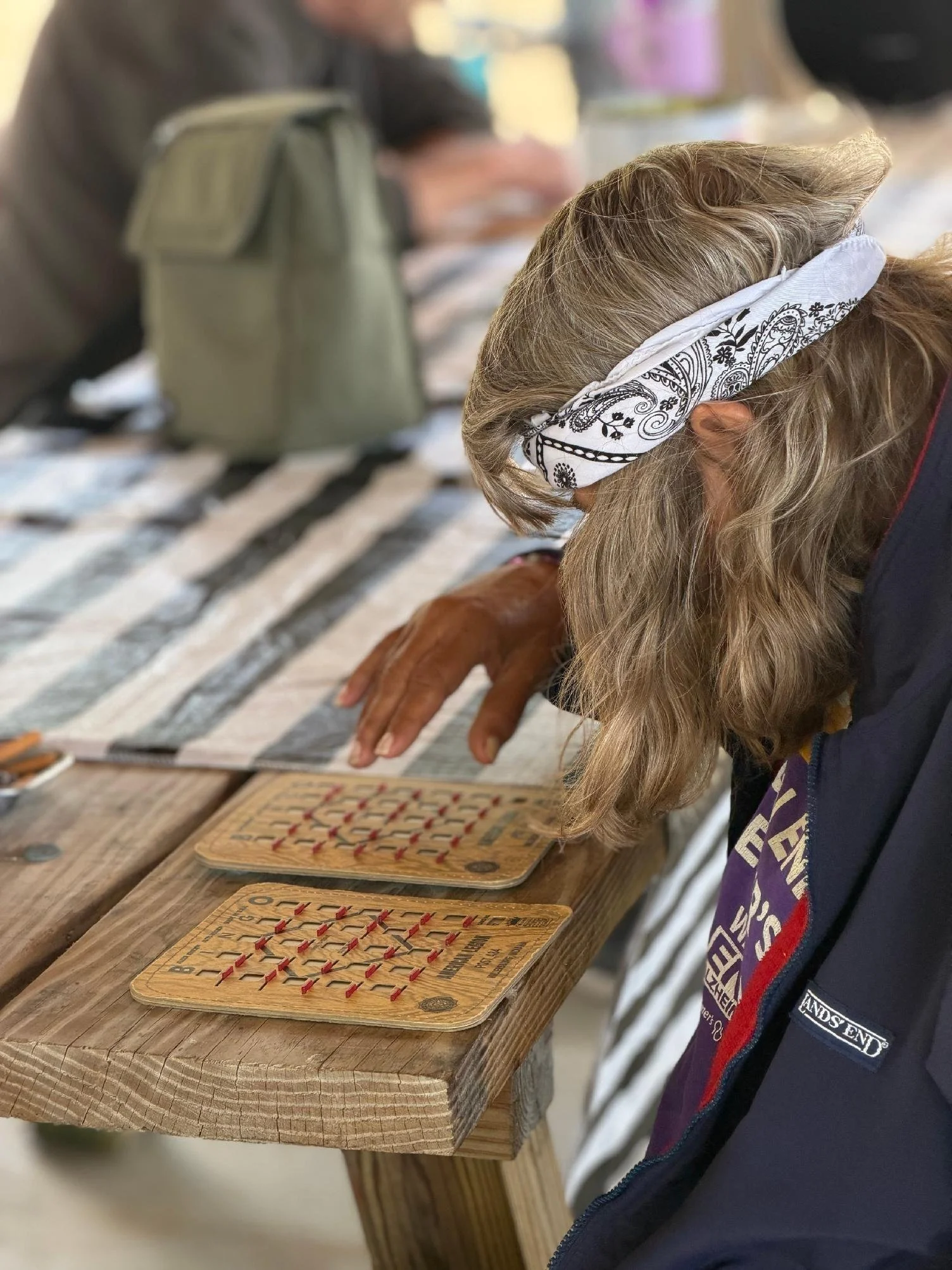 A person with long blonde hair wearing a white bandana and a purple shirt plays a vintage wooden game at a rustic wooden table, with other people and games visible in the background.