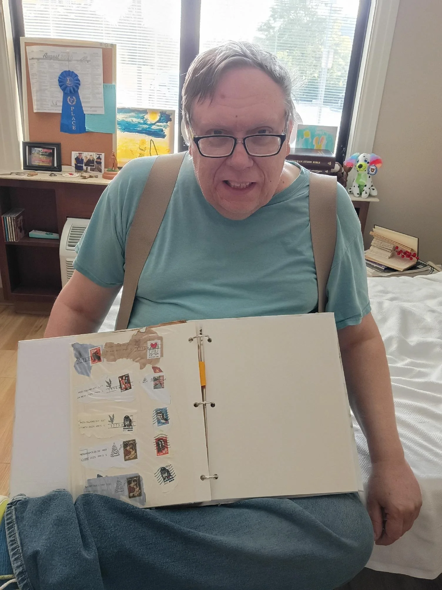 A man with glasses and light hair sitting on a bed, looking at a scrapbook with various stamps and postcards. Behind him, a window with blinds, a bulletin board with a blue ribbon, and some artwork and toys on the windowsill are visible.