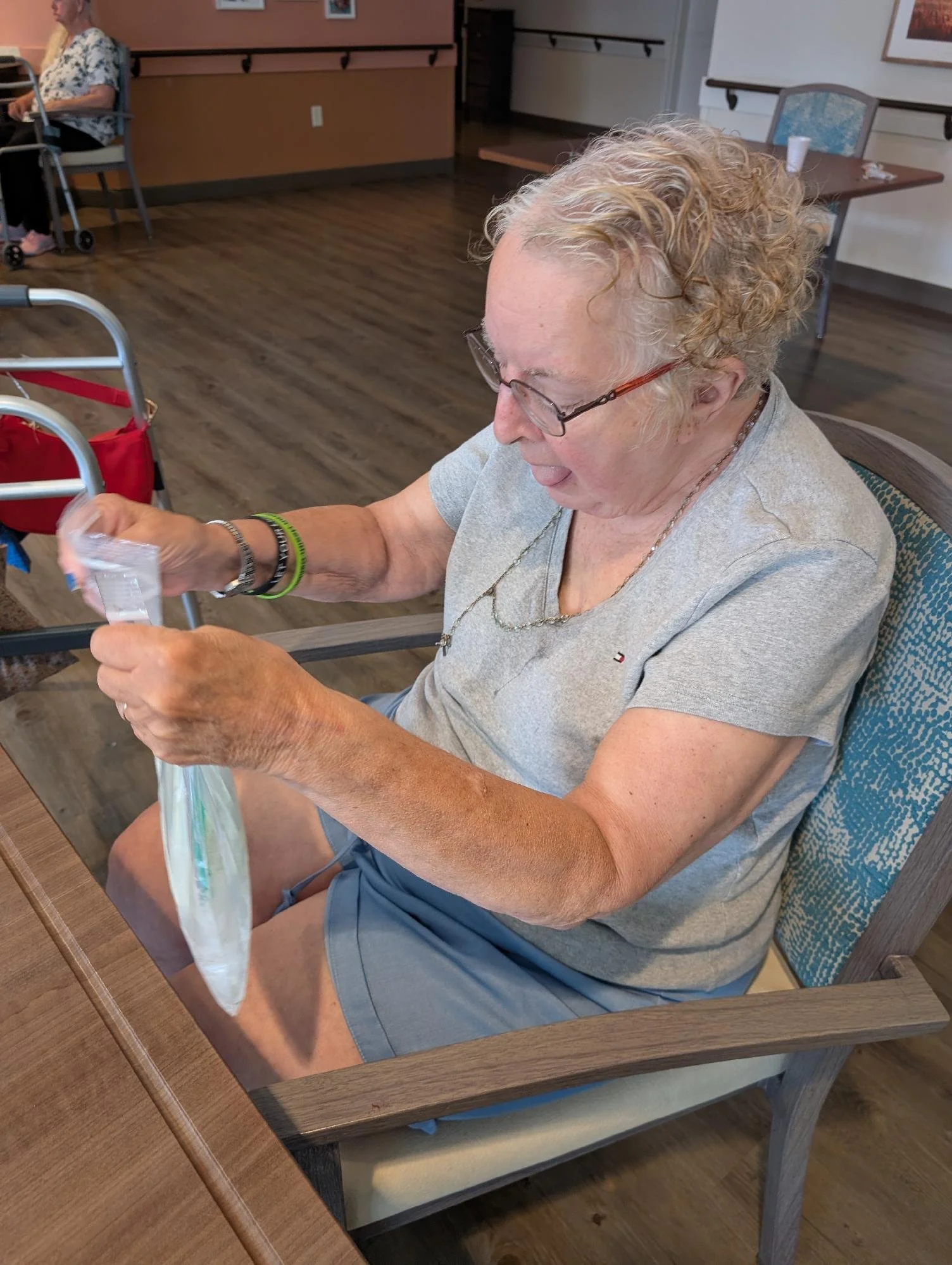 An elderly woman with curly blonde hair and glasses sitting at a wooden table in a dining area, opening a plastic bag. There is a walker beside her and another woman sitting in the background.
