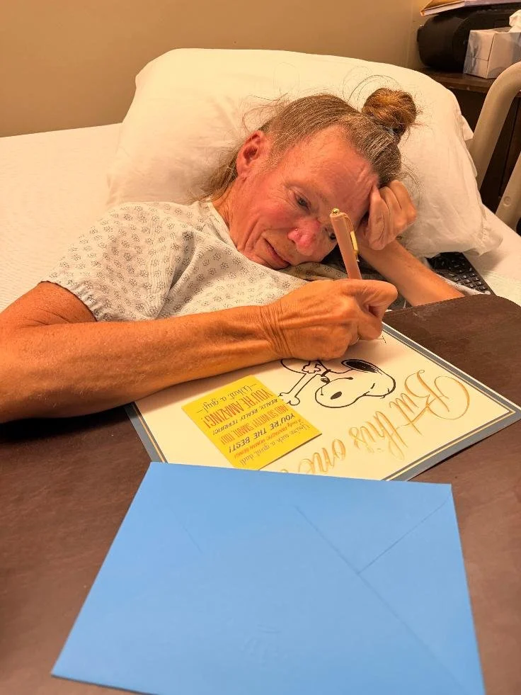 An elderly woman lying in bed, smiling, and signing a certificate with a pink pen. She has gray hair tied back and is wearing a hospital gown. The certificate features Snoopy and text.”}