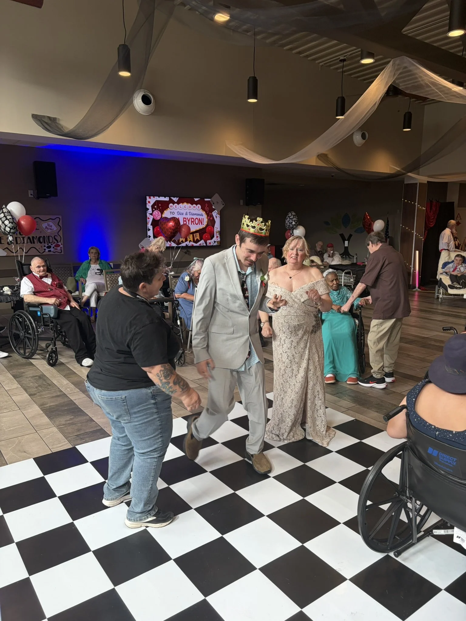 Celebrants and guests celebrating at an indoor fantasy-themed event, with people dancing on a black and white checkered dance floor, decorated with balloons, a crown, and a large screen with a welcome message.