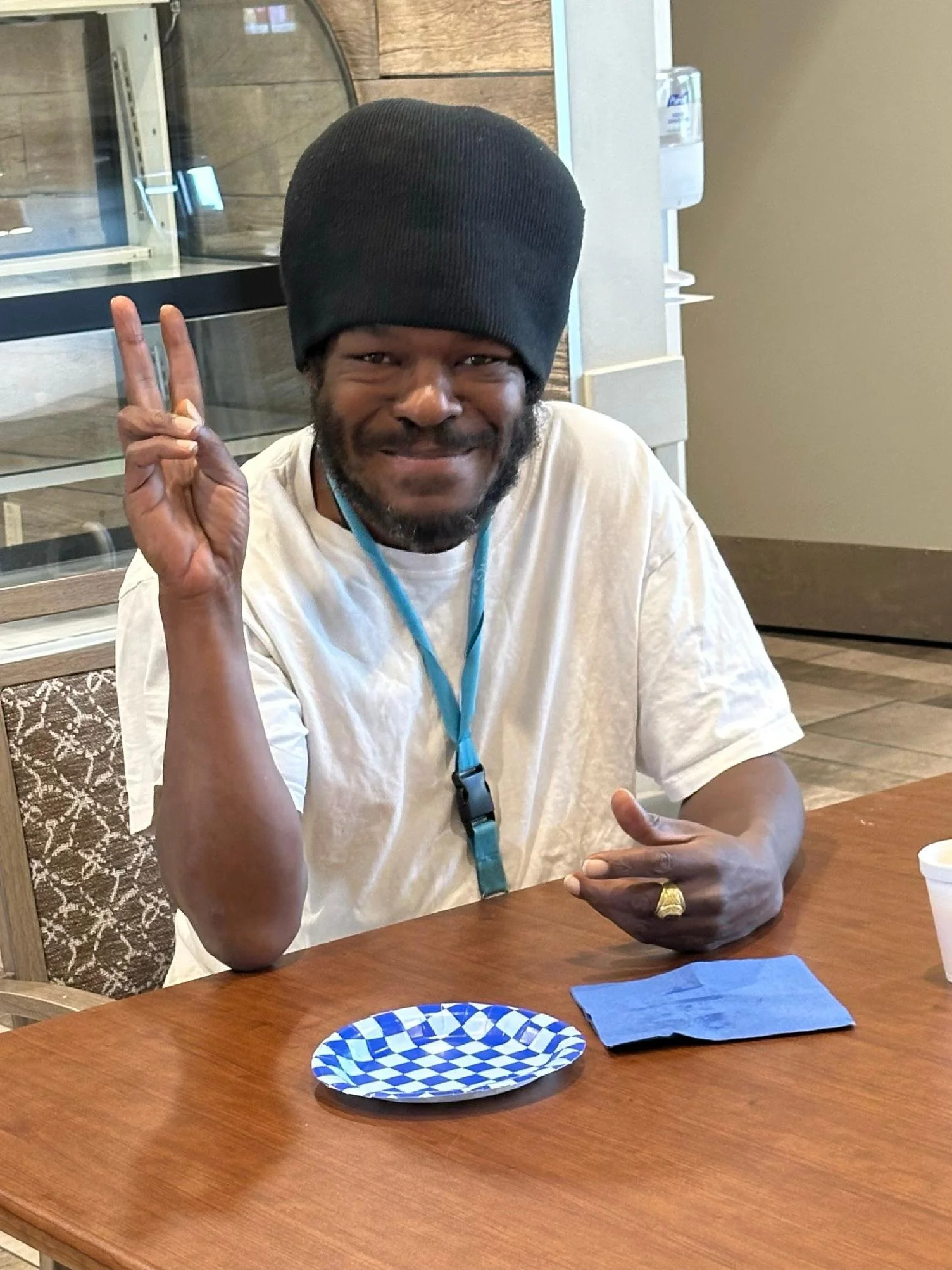A man wearing a black beanie and a white t-shirt sitting at a wooden table, making a peace sign with his right hand and smiling at the camera. On the table, there is a blue and white checkered plate, a folded blue napkin, and a white cup.