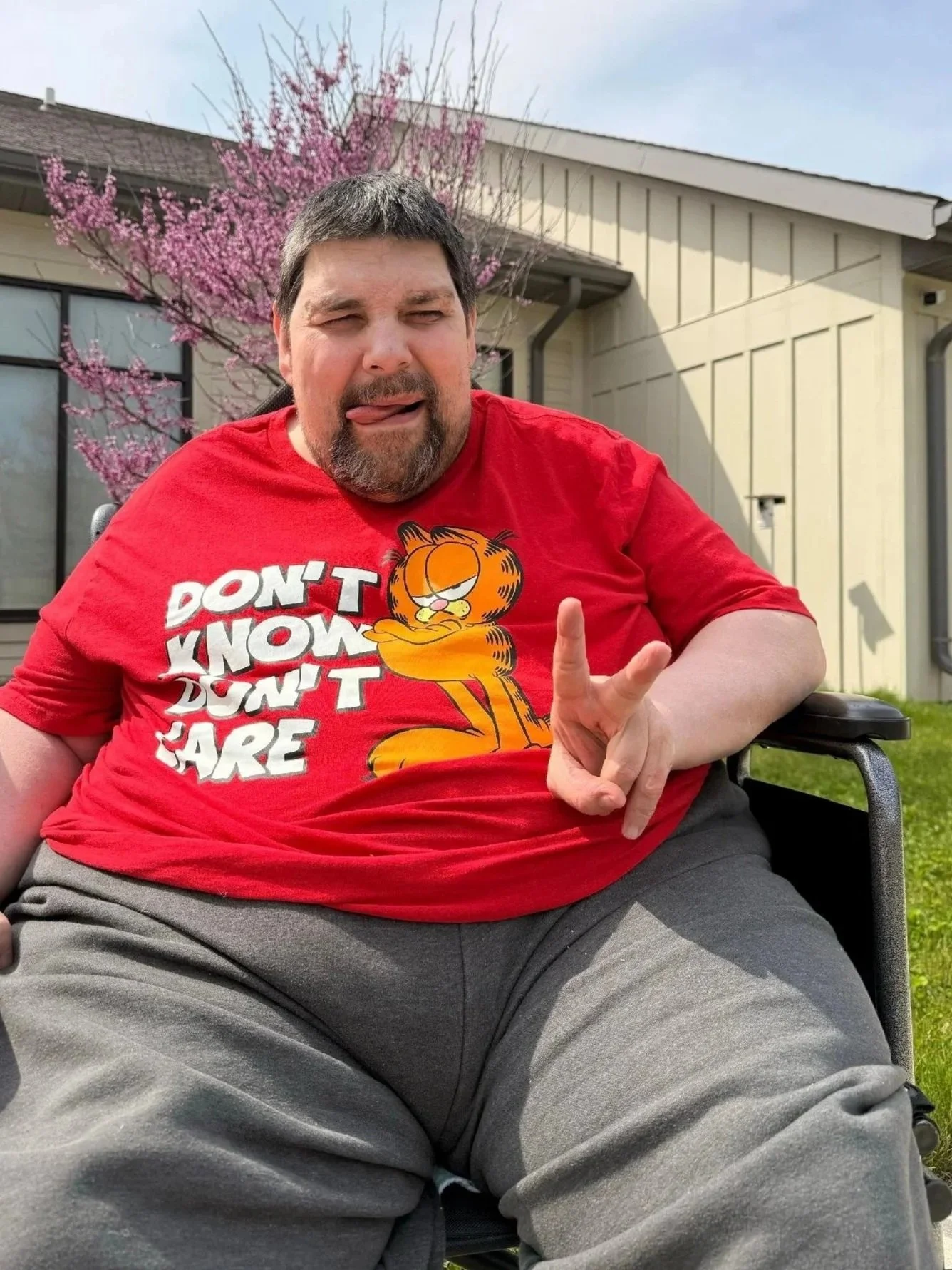 A man wearing a red Garfield T-shirt with the phrase 'Don't Know Don't Care' showing a peace sign, sitting outdoors with a house, a pink flowering tree, and a lawn in the background.
