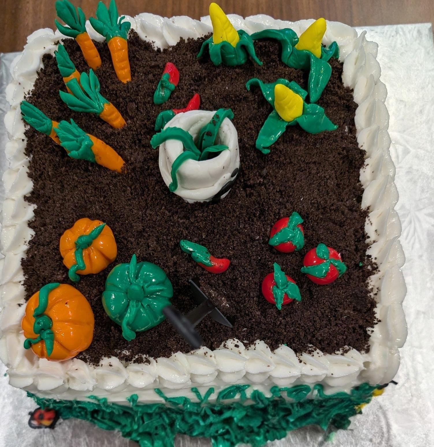 A square cake decorated as a vegetable garden with colorful plastic vegetables and soil made from crushed cookies.