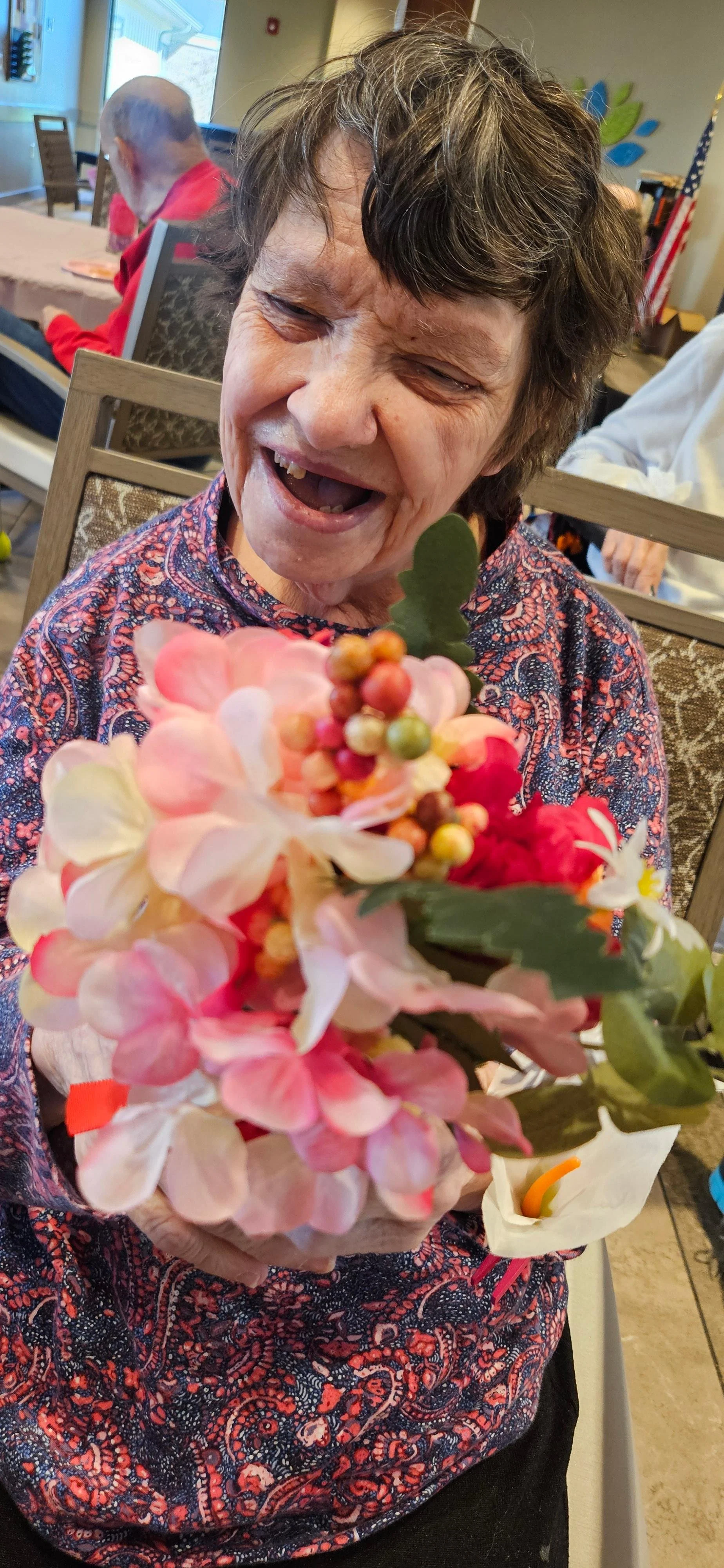 An elderly woman with short gray hair, wearing a patterned shirt, smiling and holding a bouquet of pink, white, and red flowers with green leaves.